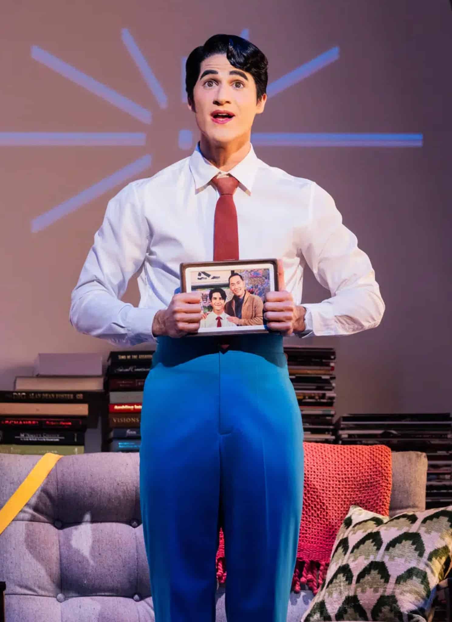 A person in blue pants, white shirt, and red tie stands onstage holding a framed photo, with books, a chair, a lamp, and a potted plant in the background. Blue lines radiate on the wall behind them.