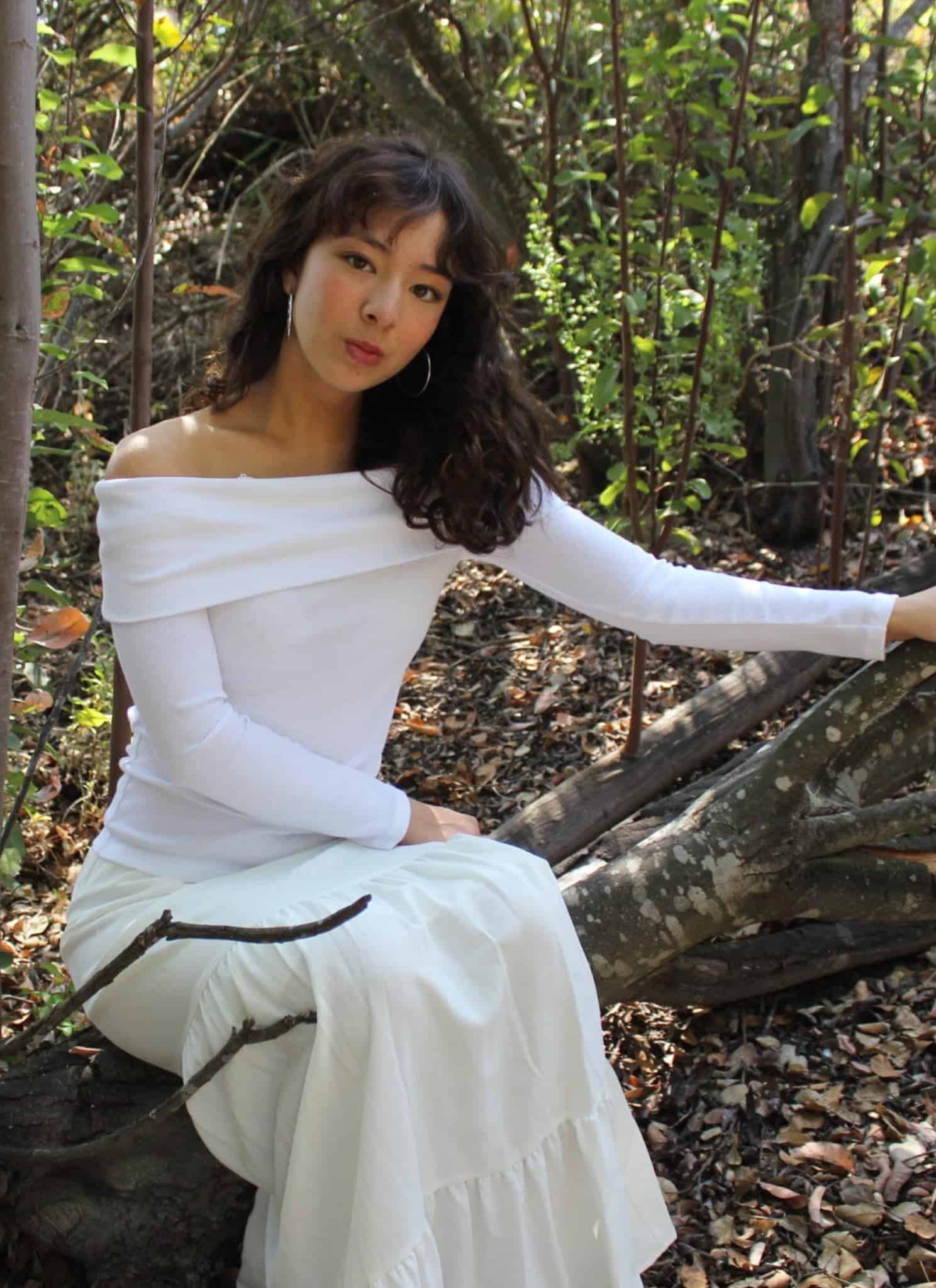 A woman in a long white off-the-shoulder dress sits on a tree branch in a forest, surrounded by trees and fallen leaves, looking thoughtfully at the camera.