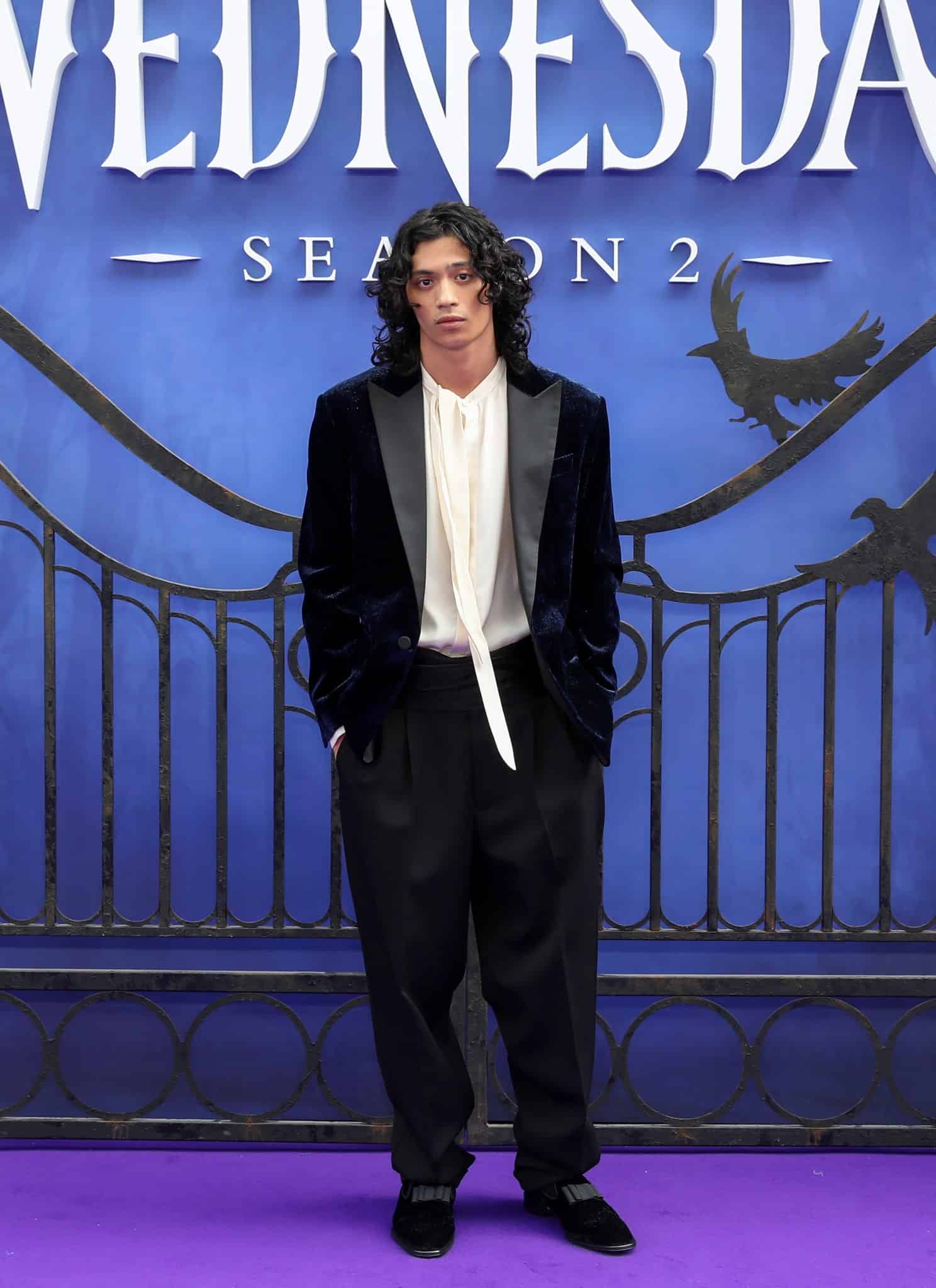 A person with long, curly dark hair wearing a black velvet jacket, white shirt, and black pants stands in front of a blue backdrop with Wednesday Season 2 and a wrought iron gate design.