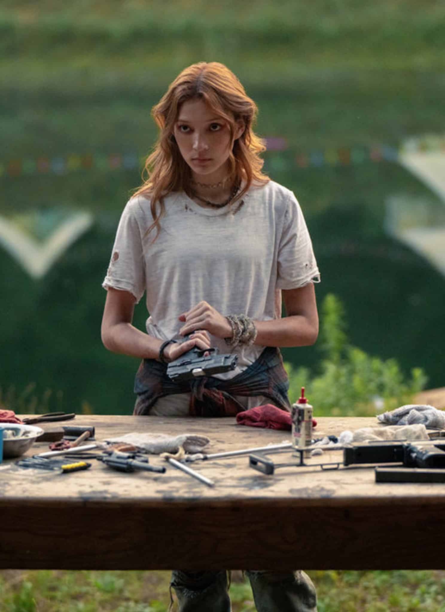 A young woman stands behind a workbench covered with tools and gun parts near a lake, wearing a white t-shirt with bracelets on her wrists. Colorful pennant flags hang nearby, and trees are reflected in the water.
