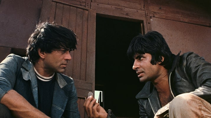 Two men with dark hair sit close together outdoors, one holding a coin and speaking seriously to the other. They are wearing casual jackets, and a wooden structure is visible in the background.