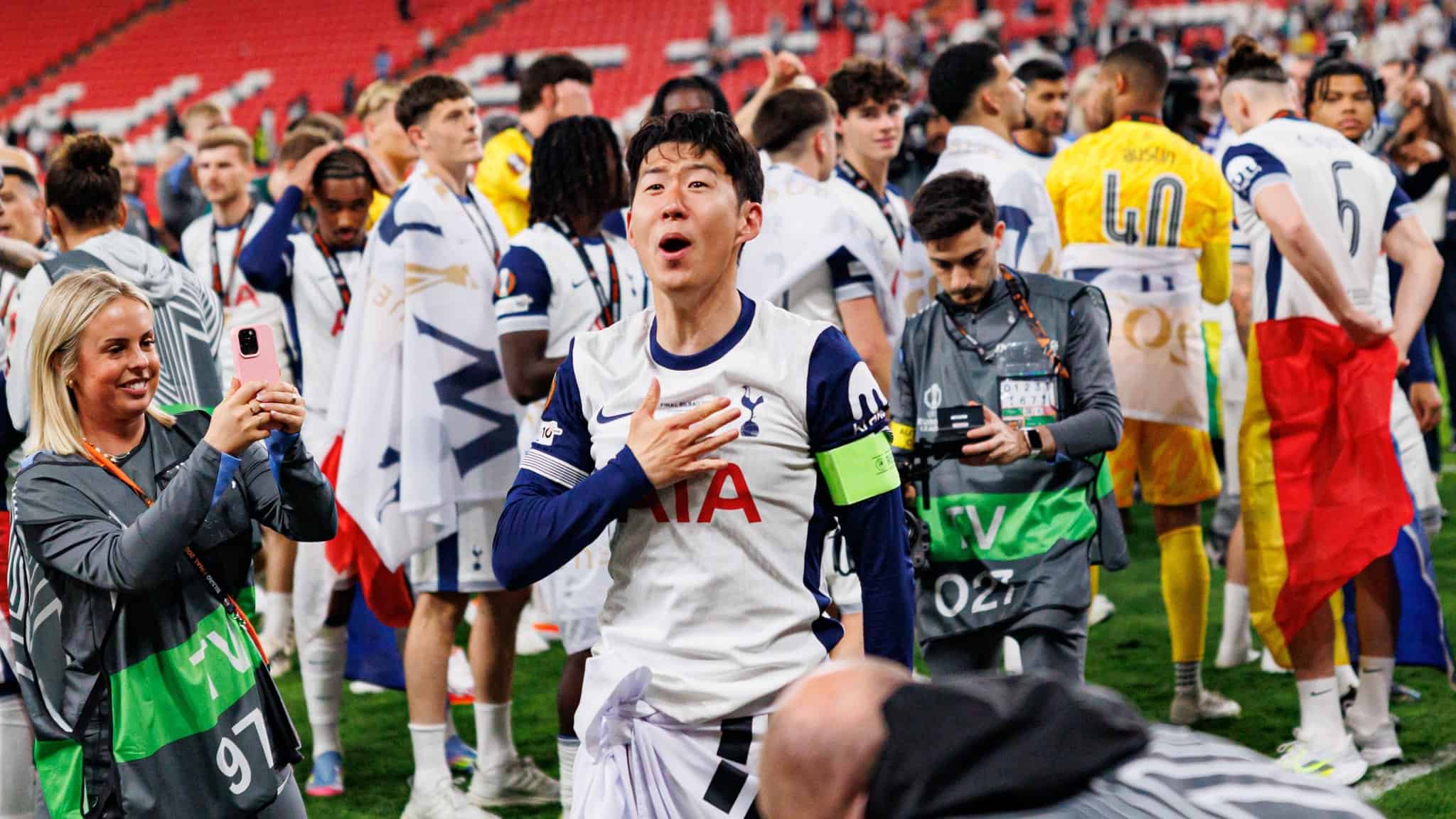 Tottenham Hotspur soccer player Son Heung-min, wearing a captains armband, smiles and gestures with his hand on his chest as fans and media take photos and players gather in the background on the field.
