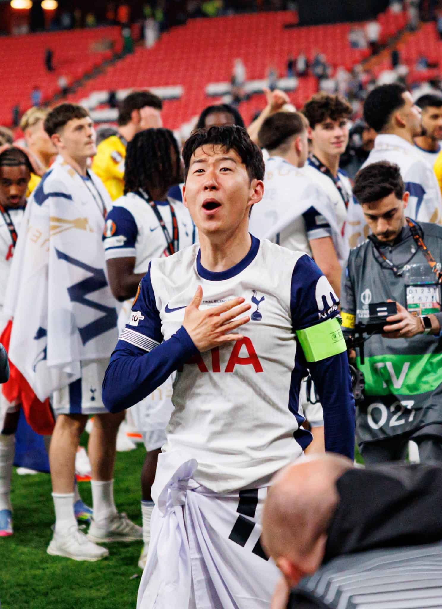 Tottenham Hotspur soccer player Son Heung-min, wearing a captains armband, smiles and gestures with his hand on his chest as fans and media take photos and players gather in the background on the field.