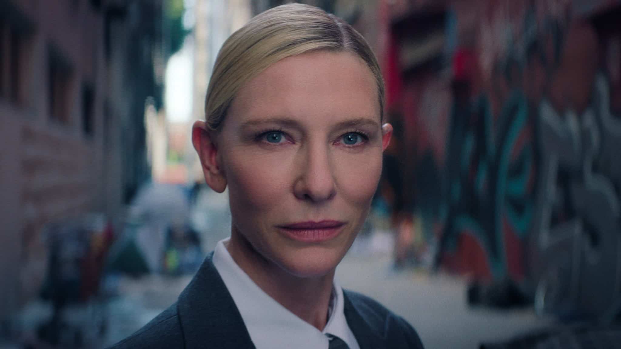 A person with blonde hair pulled back, wearing a suit and tie, stands in an urban alleyway with graffiti-covered walls and blurred background. They have a serious expression and are looking directly at the camera.