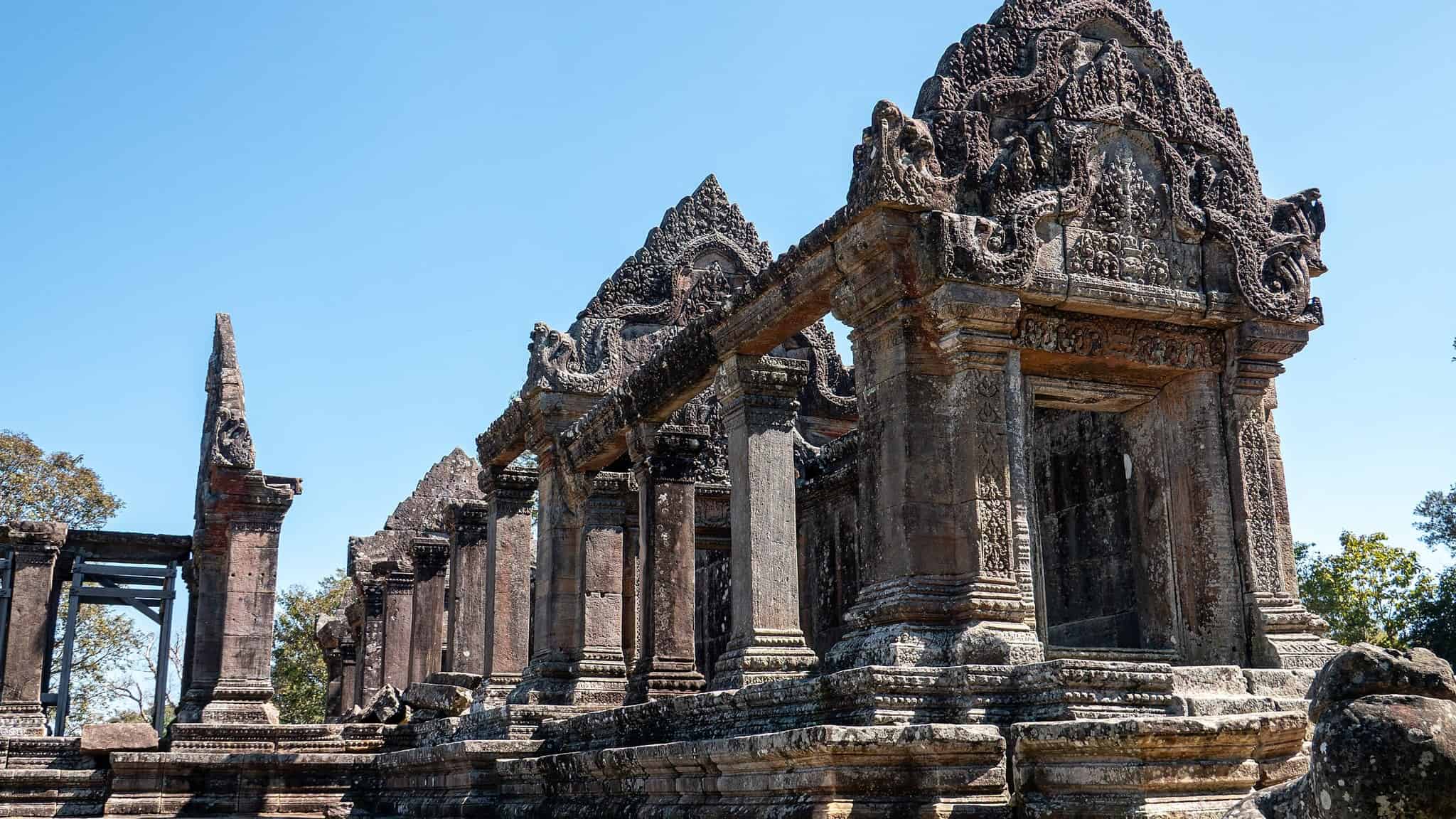Ancient stone temple ruins with intricate carvings and tall columns, set against a clear blue sky and surrounded by some trees.