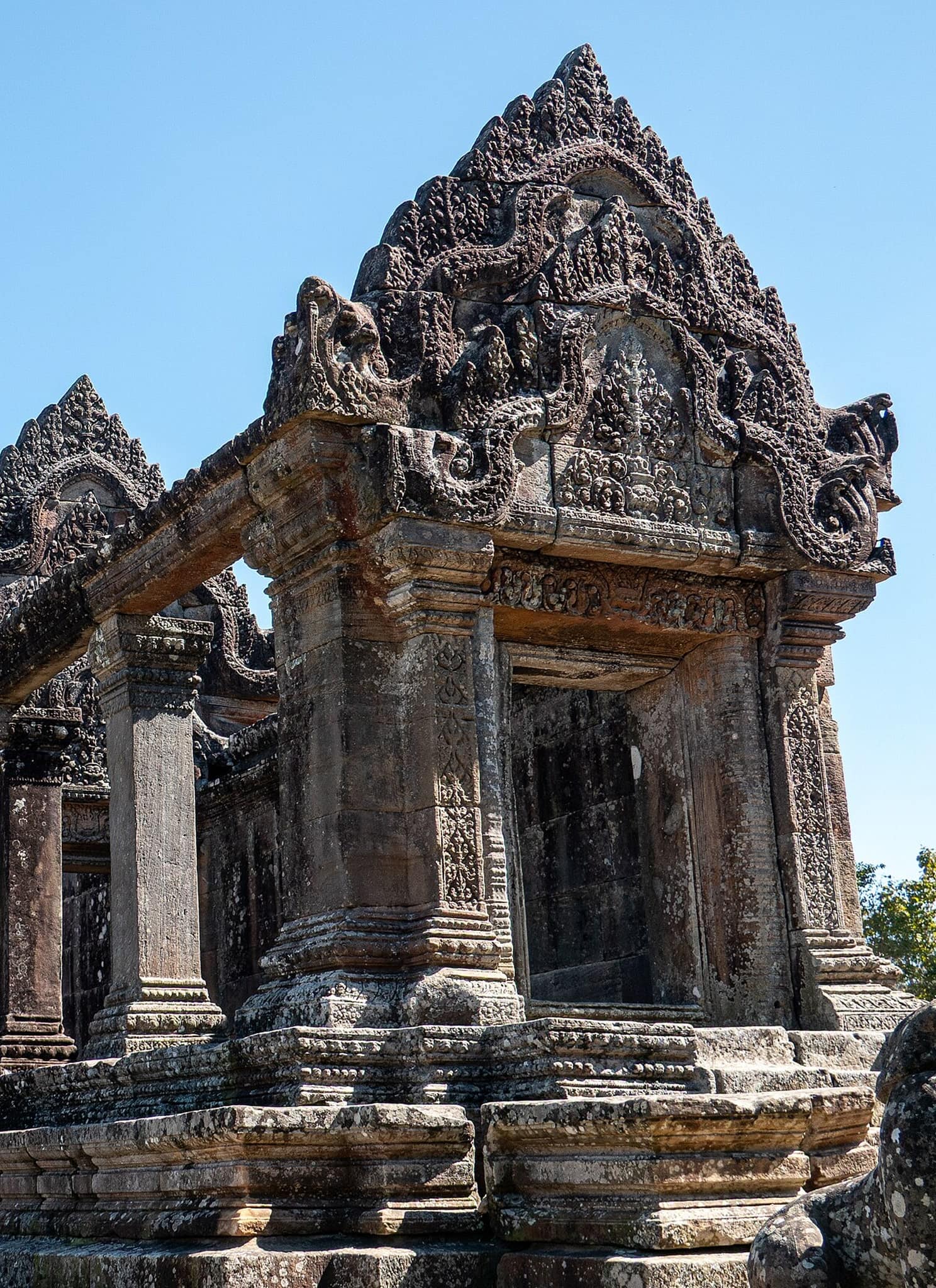 Ancient stone temple ruins with intricate carvings and tall columns, set against a clear blue sky and surrounded by some trees.