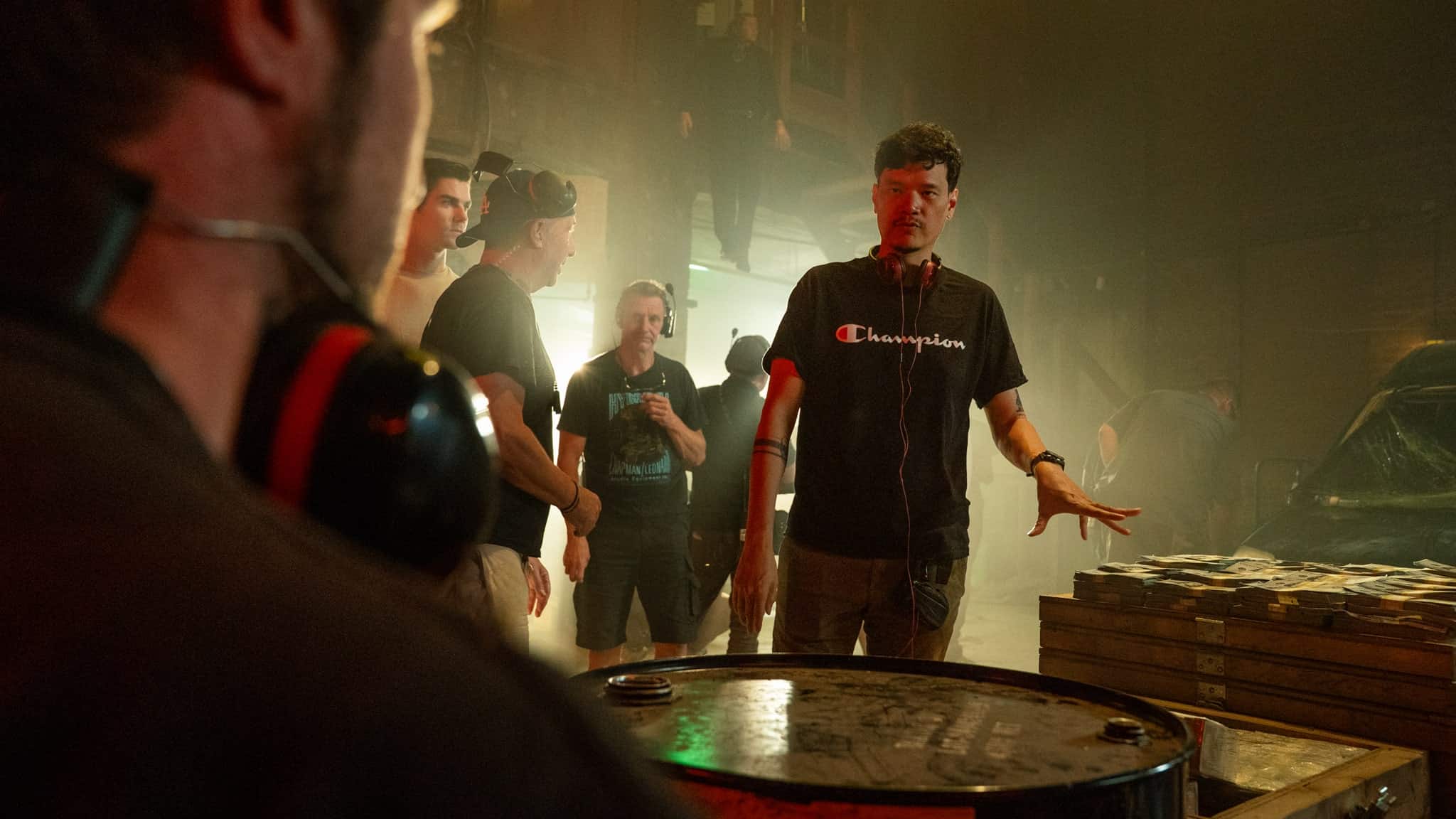 A man wearing headphones and a Champion shirt gestures while talking to a group of people in a dim, industrial setting with a metal barrel and stacks of items nearby.
