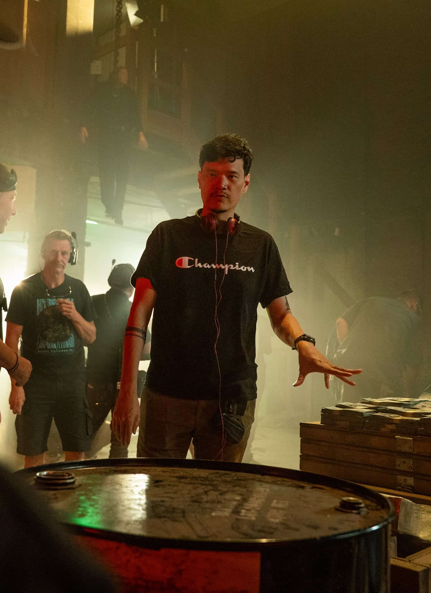 A man wearing headphones and a Champion shirt gestures while talking to a group of people in a dim, industrial setting with a metal barrel and stacks of items nearby.