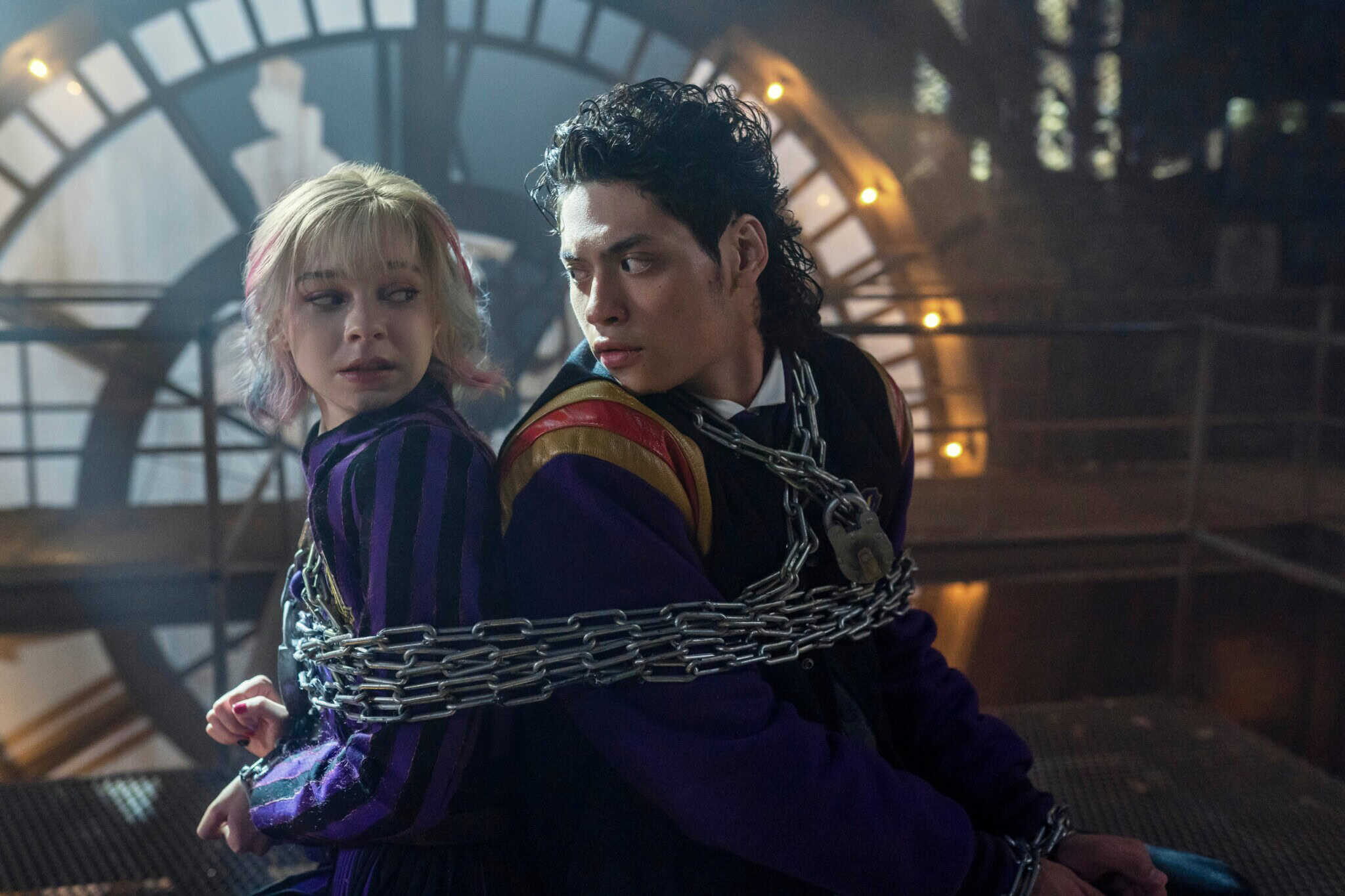 Two young people, a blonde girl with pink highlights and a dark-haired boy in school uniforms, sit back-to-back tied with chains. They appear worried, inside a building with a large clock face behind them.