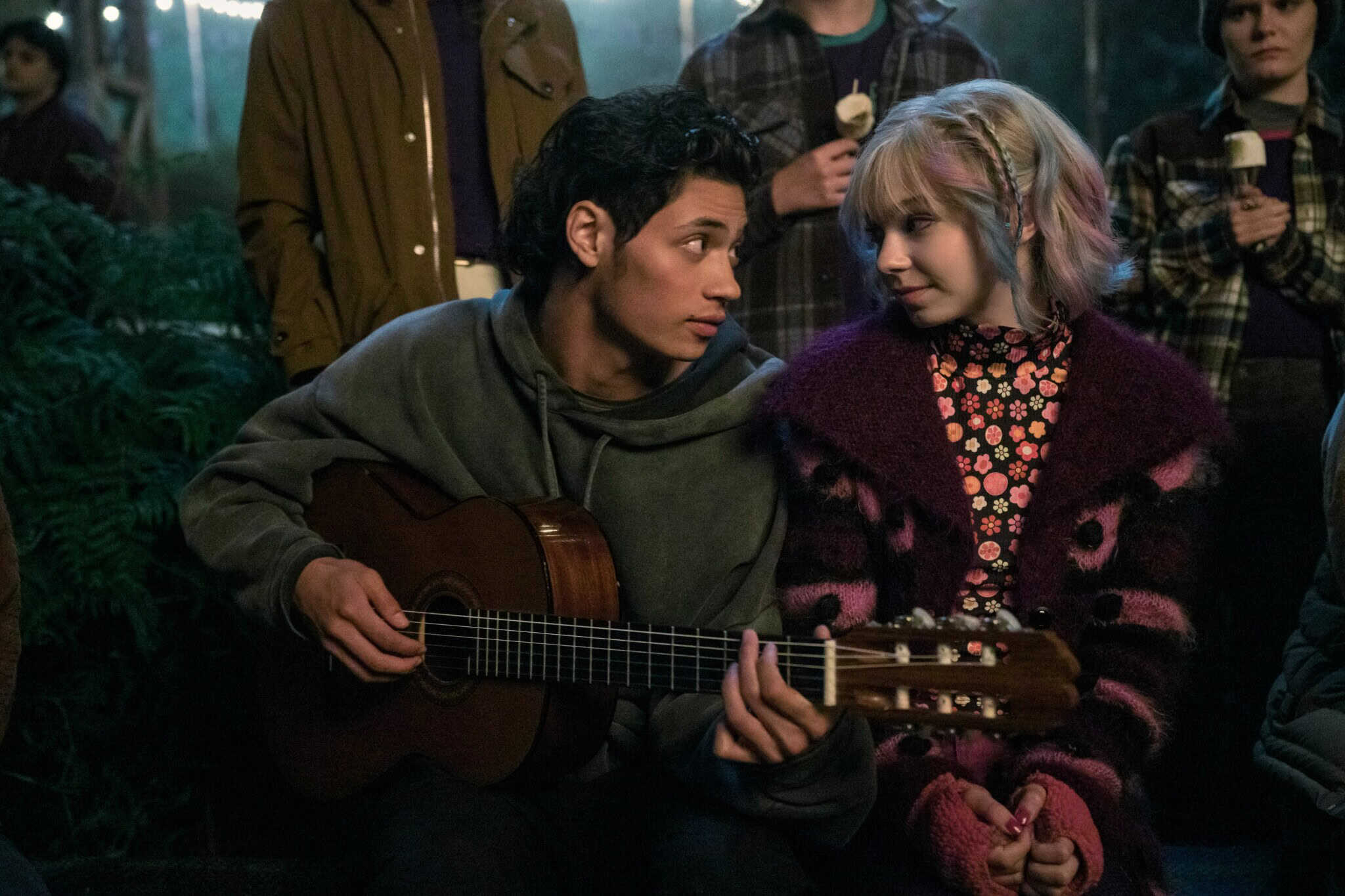 A young man plays guitar and gazes at a young woman with pastel hair and a purple sweater. They sit close together, surrounded by people at an outdoor nighttime gathering.