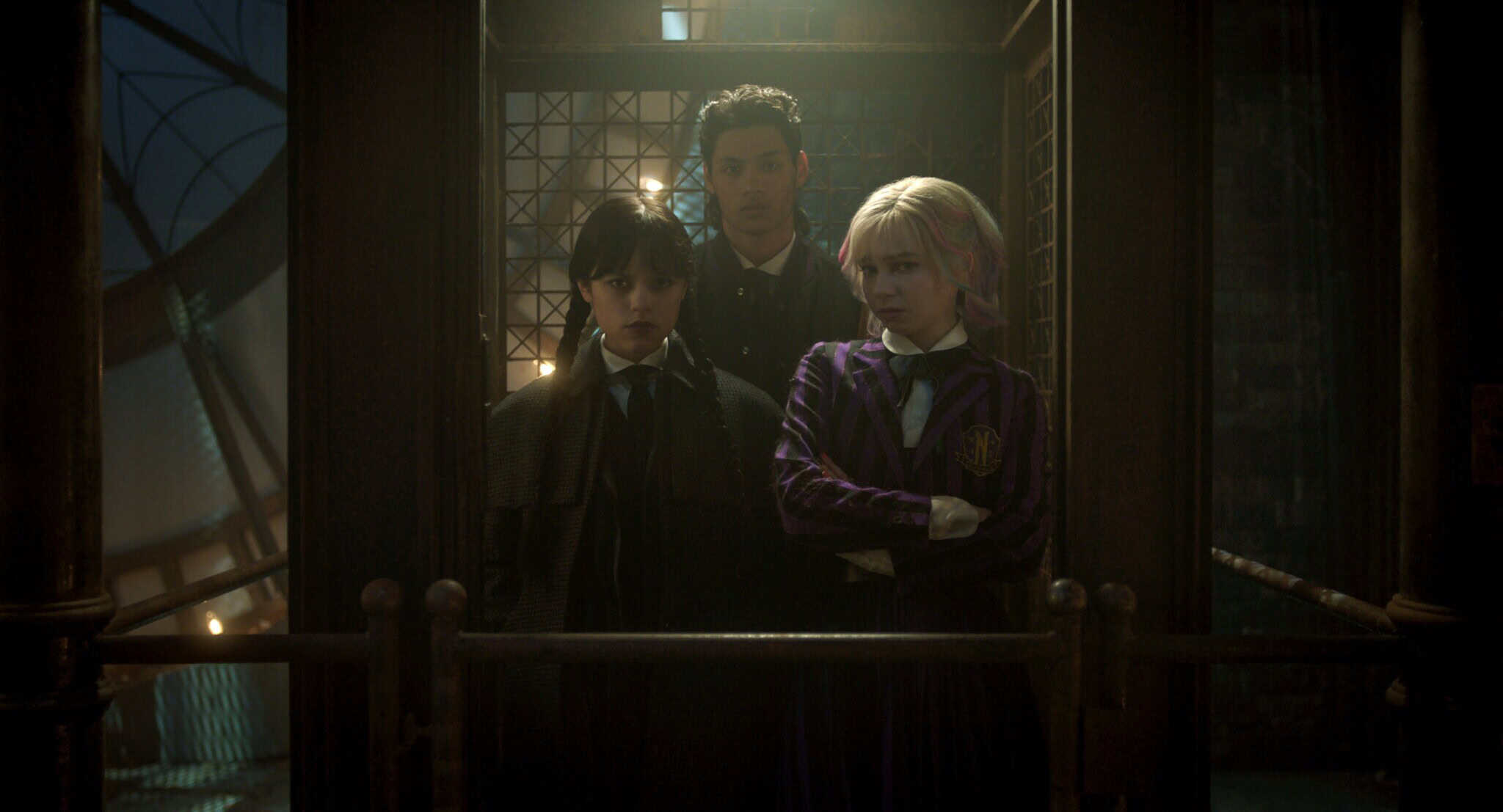 Three teenagers stand in an old-fashioned elevator with metal lattice doors. Two girls, one with dark hair and one with light hair wearing a purple blazer, are in front, while a boy stands behind them. The scene is dimly lit.