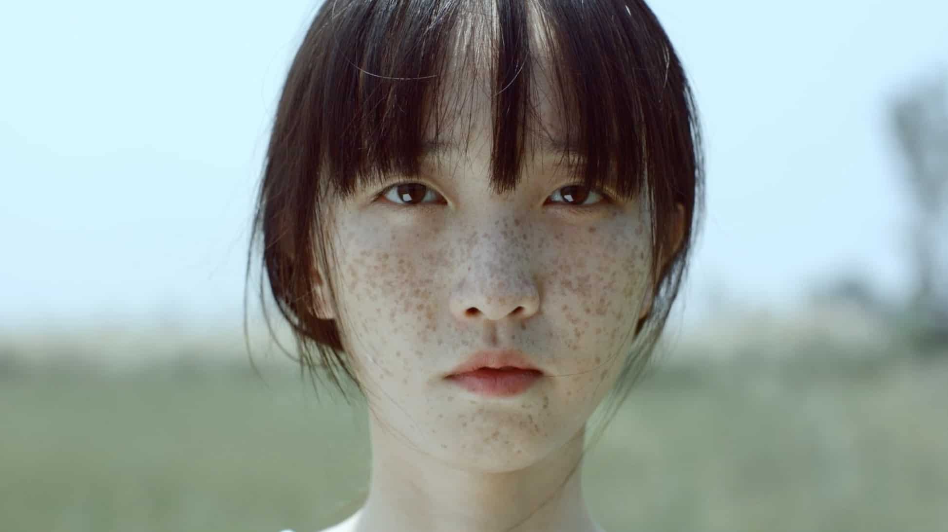 A young person with straight dark hair and freckles looks directly at the camera, standing outdoors with a blurred grassy background under bright daylight.