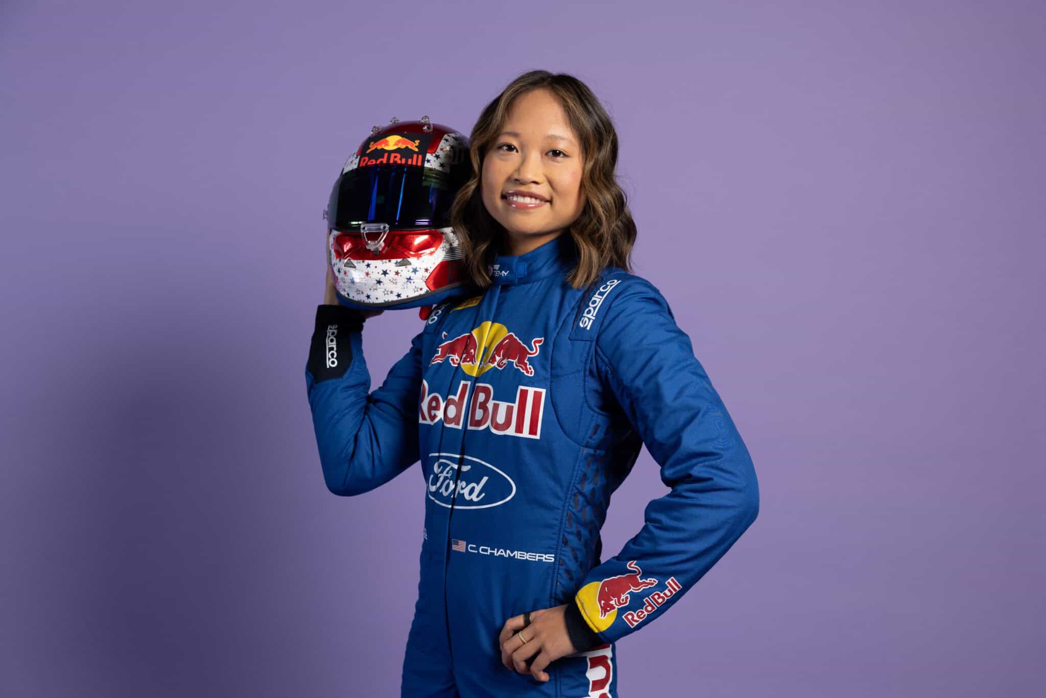 A woman in a blue racing suit with Red Bull and Ford logos holds a decorated racing helmet next to her face, standing against a plain purple background and smiling confidently.