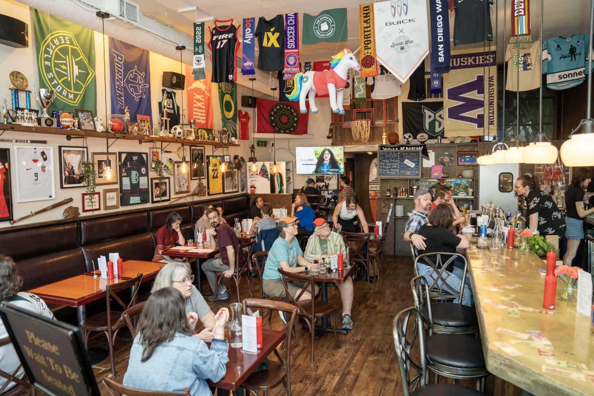A lively restaurant or bar with people dining and chatting. Sports flags, jerseys, and memorabilia cover the walls, and a large stuffed unicorn hangs from the ceiling. The atmosphere is casual and vibrant.