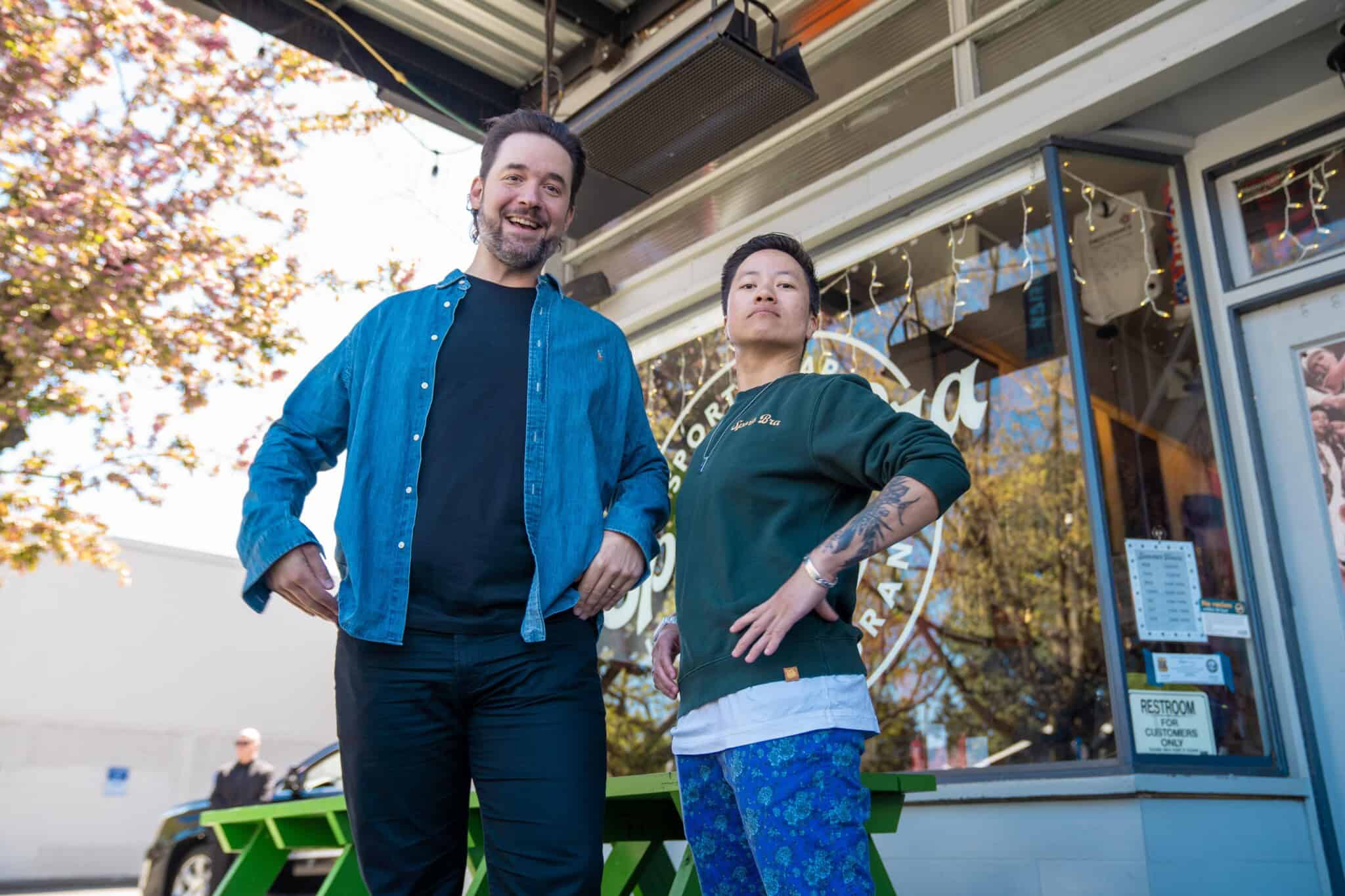 Two people stand confidently outside a restaurant with large windows and visible signage. One wears a blue shirt over a black tee, the other has tattoos and wears a white shirt and blue pants. Trees and a green bench are nearby.
