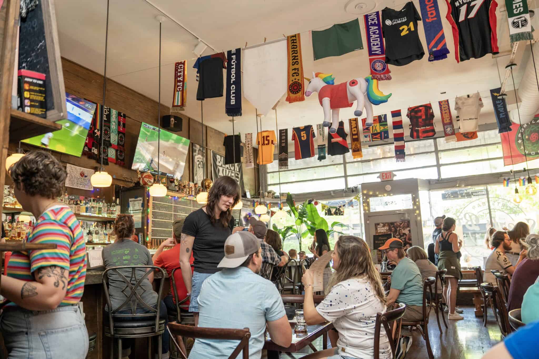 A lively bar scene with people socializing at tables; soccer scarves, jerseys, and flags hang from the ceiling, while TVs show sports. Sunlight streams through large windows in the background.