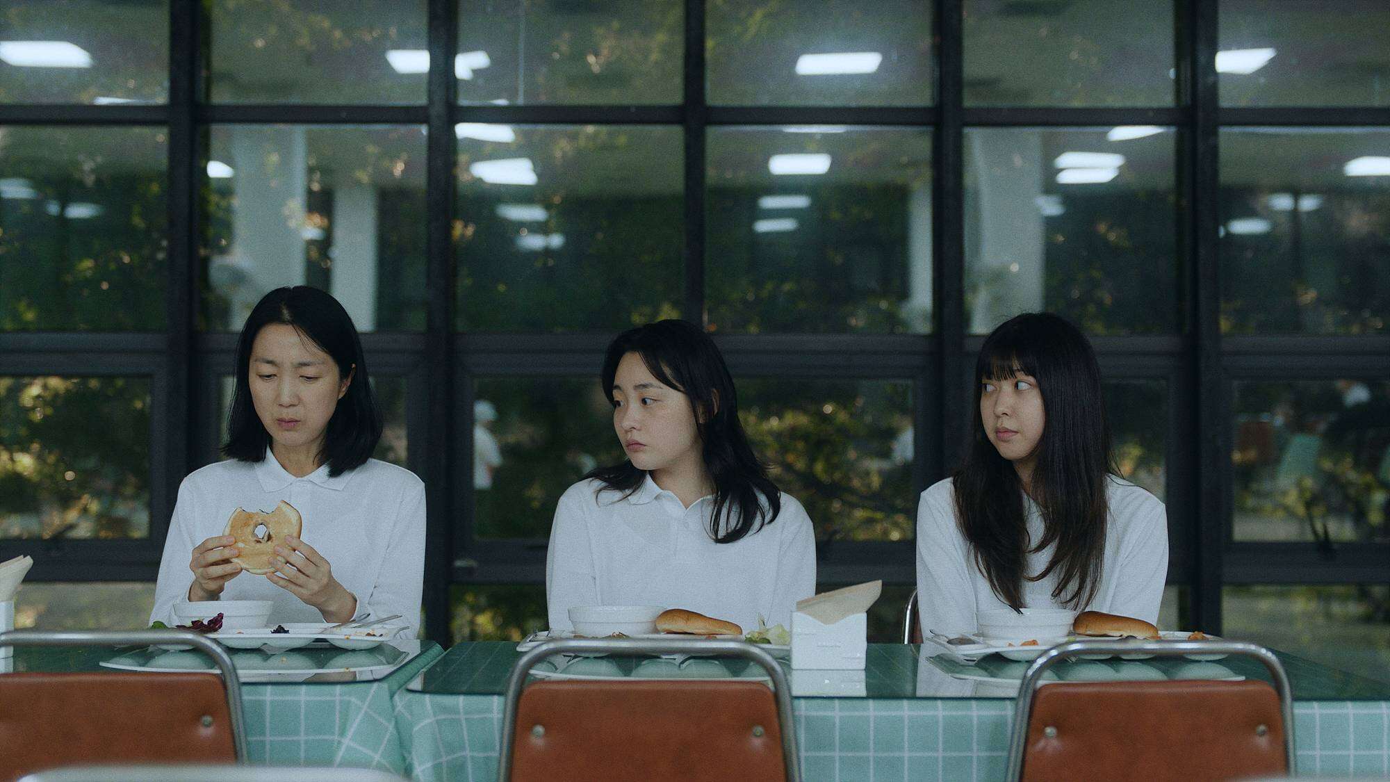 Three women in white shirts sit at a table with food in a cafeteria. The woman on the left holds a sandwich, while the other two look at her. Large windows with a leafy view are in the background.