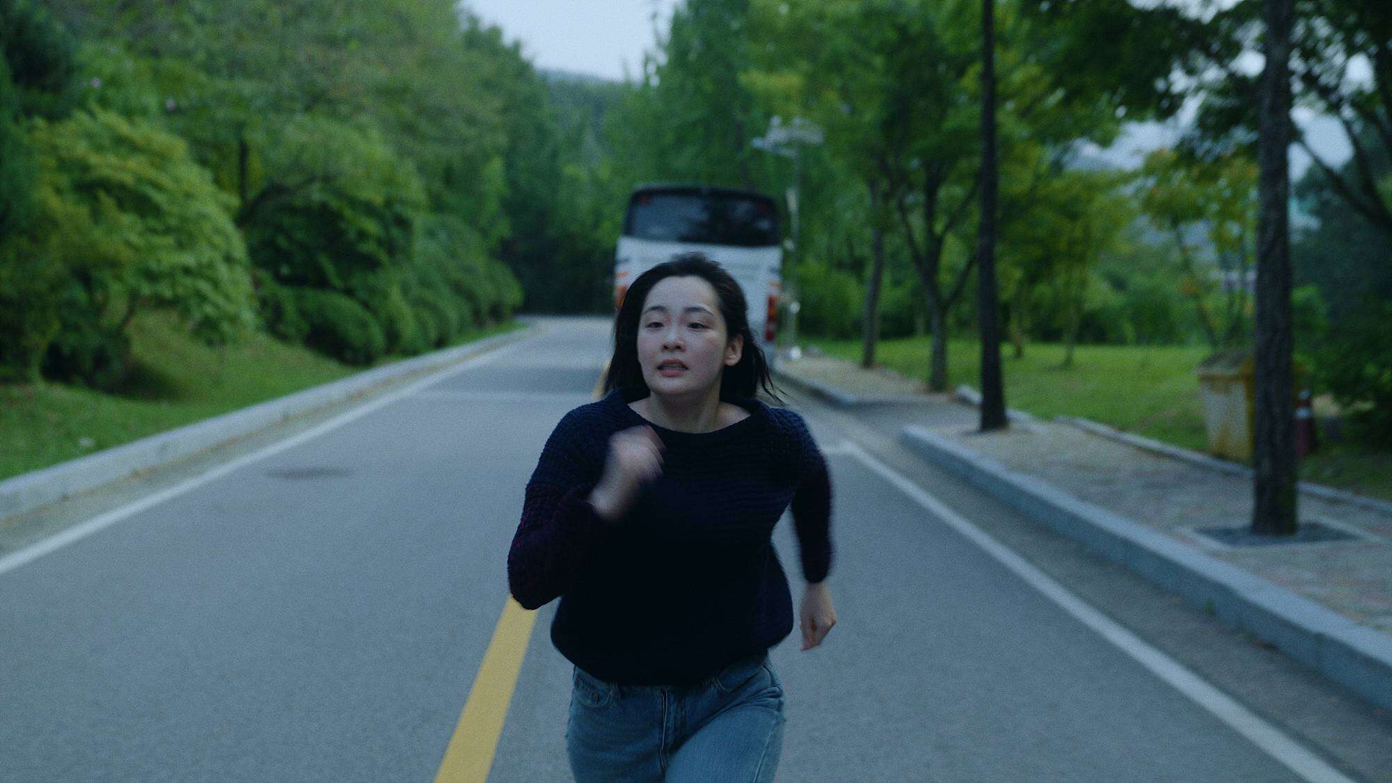 A woman in a dark sweater and jeans runs down a deserted road lined with trees and greenery, with a bus parked in the background.