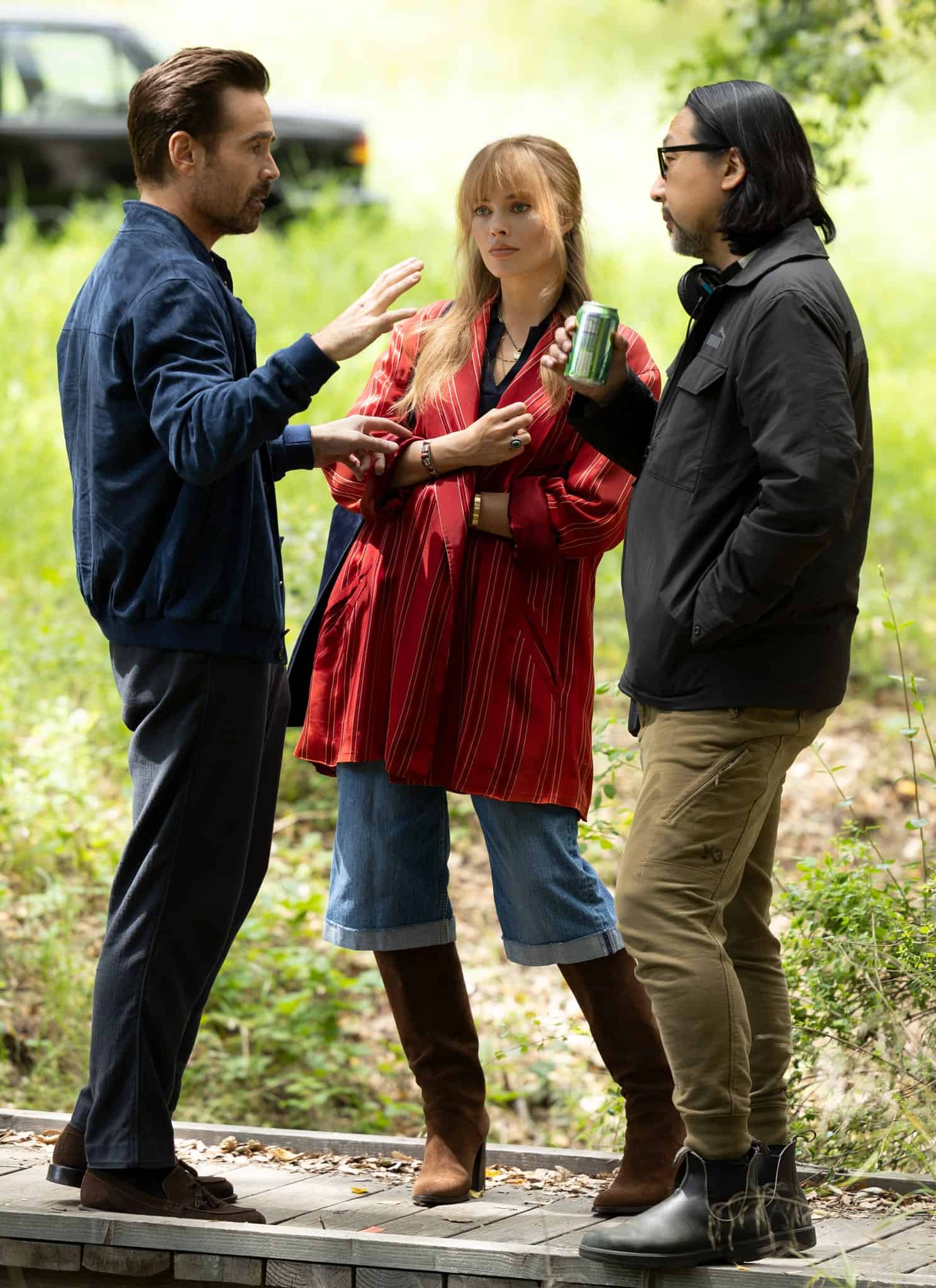 Three people stand talking in a grassy, wooded area, perhaps at the start of A Big Bold Beautiful Journey. One gestures with his hands, the woman in the center holds a can, and the third listens. A blurred black car sits among the trees in the background.