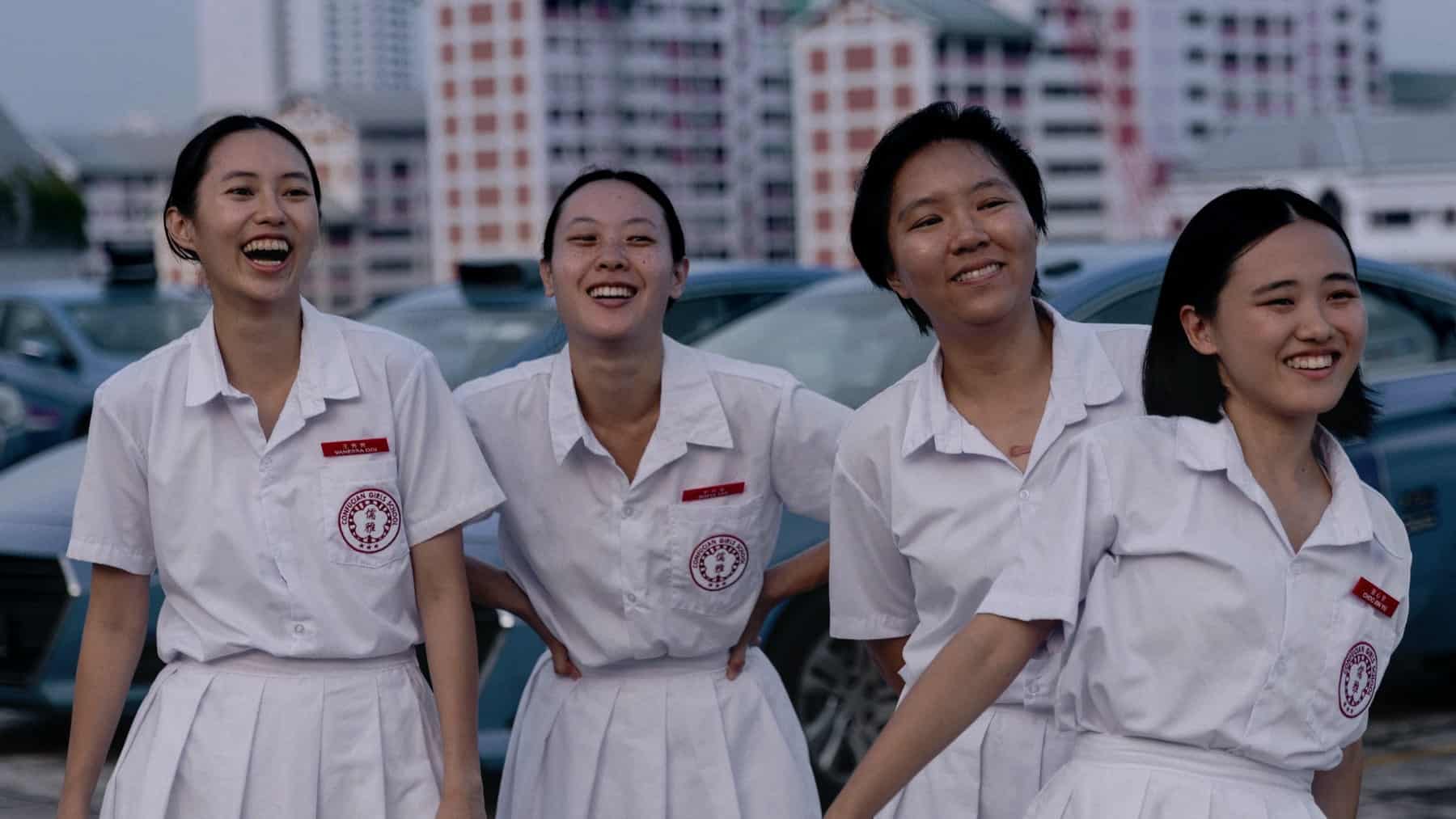 Four young women in white school uniforms with red name tags stand outside, smiling and laughing, with parked cars and tall buildings in the background.