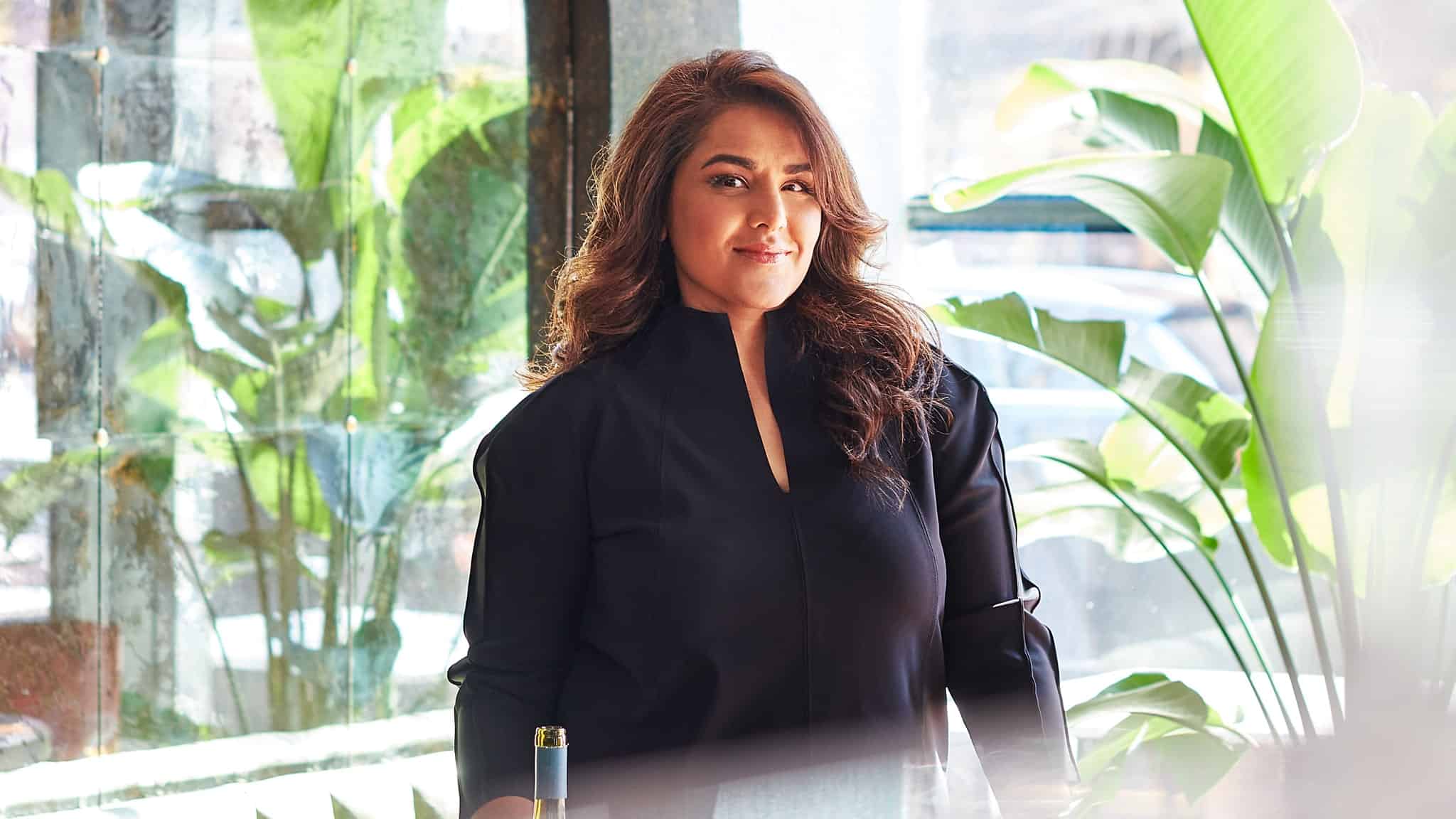 A woman with long, wavy hair wearing a black top stands indoors by large green plants, with sunlight shining through a window behind her. She is smiling softly and looking at the camera.