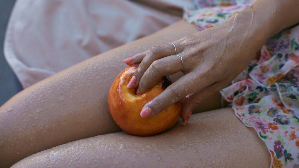 A close-up of a hand with pink nails and silver rings holding a peach on wet, bare legs. Expressing subtle sexuality, the Asian American wearer dons a floral dress as water droplets glisten on skin and fruit.