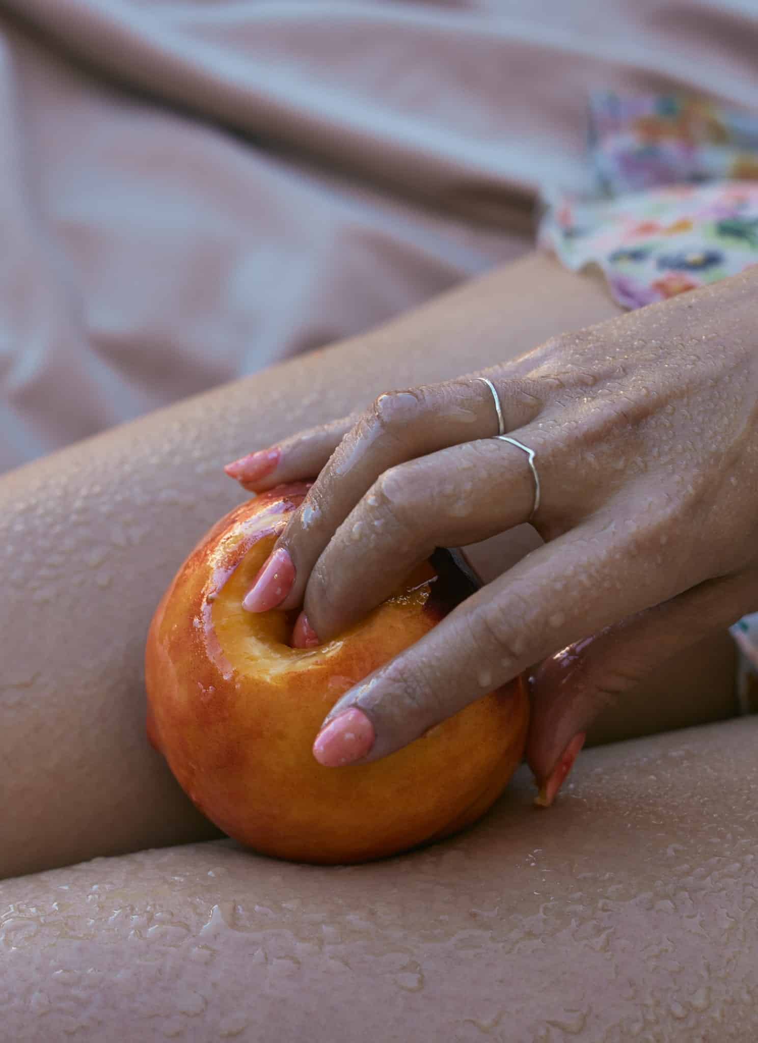 A close-up of a hand with pink nails and silver rings holding a peach on wet, bare legs. Expressing subtle sexuality, the Asian American wearer dons a floral dress as water droplets glisten on skin and fruit.