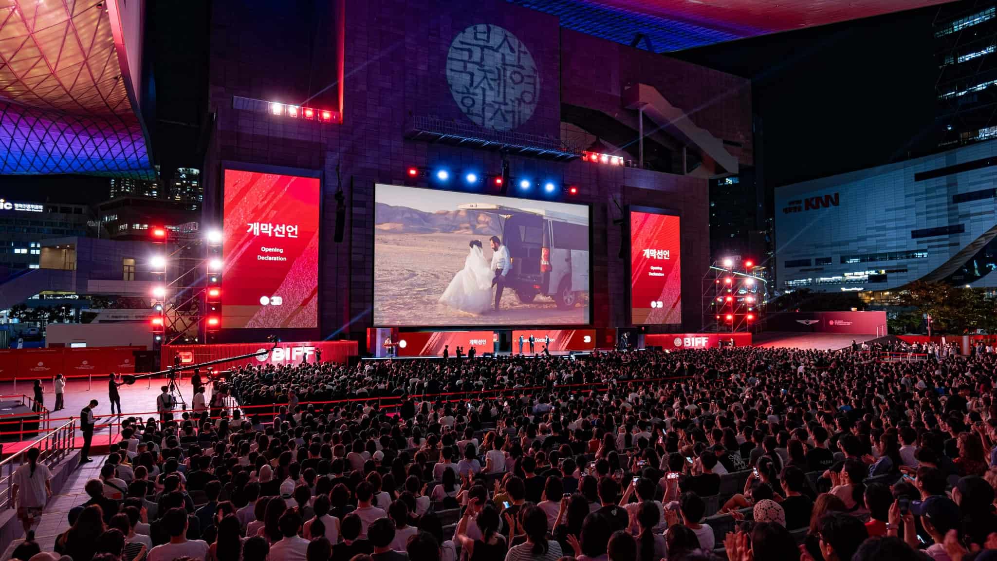 A large outdoor audience watches a film on a big screen at night during a film festival, with red lights and banners, in an urban setting.