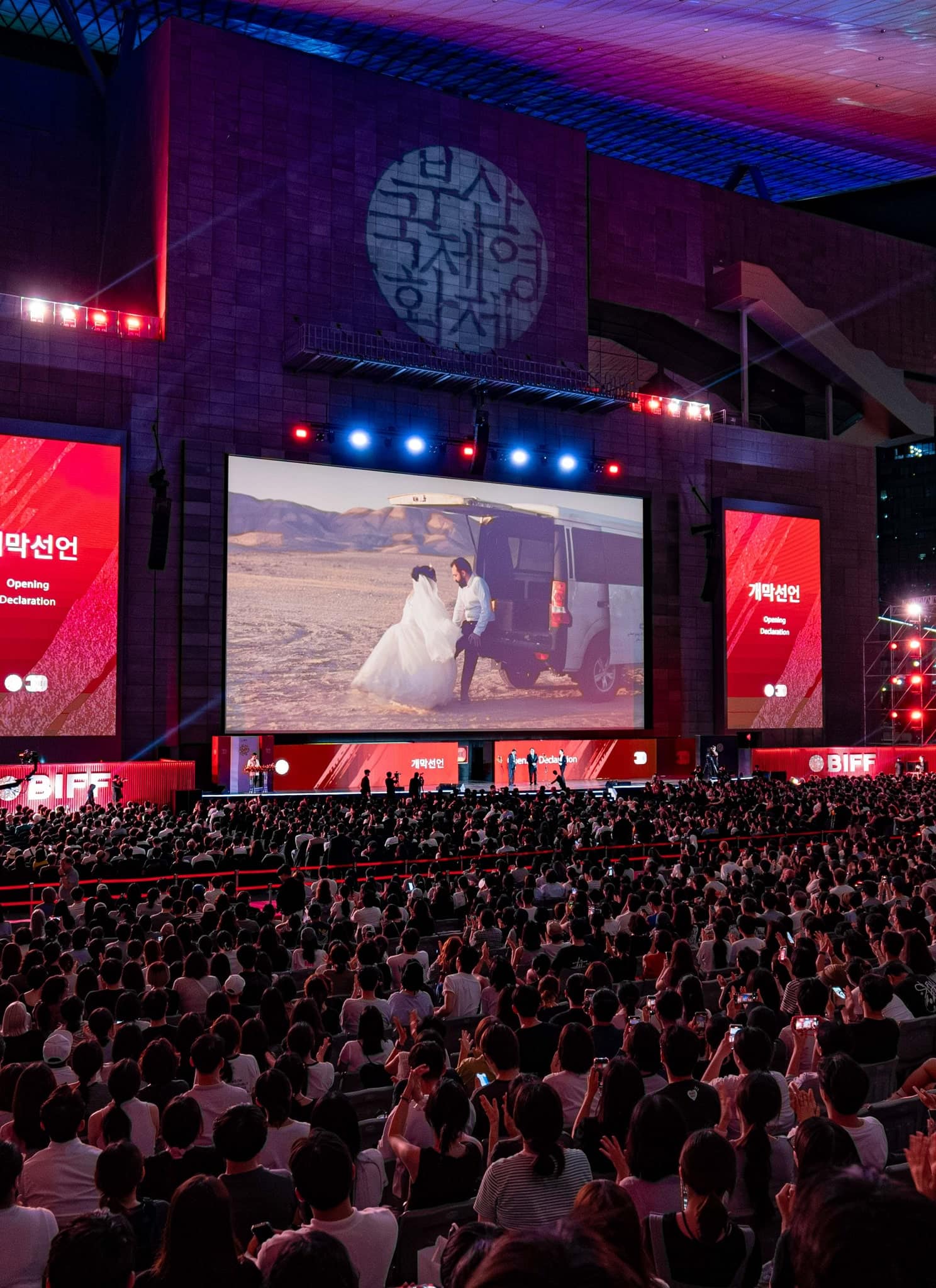 A large outdoor audience watches a film on a big screen at night during a film festival, with red lights and banners, in an urban setting.