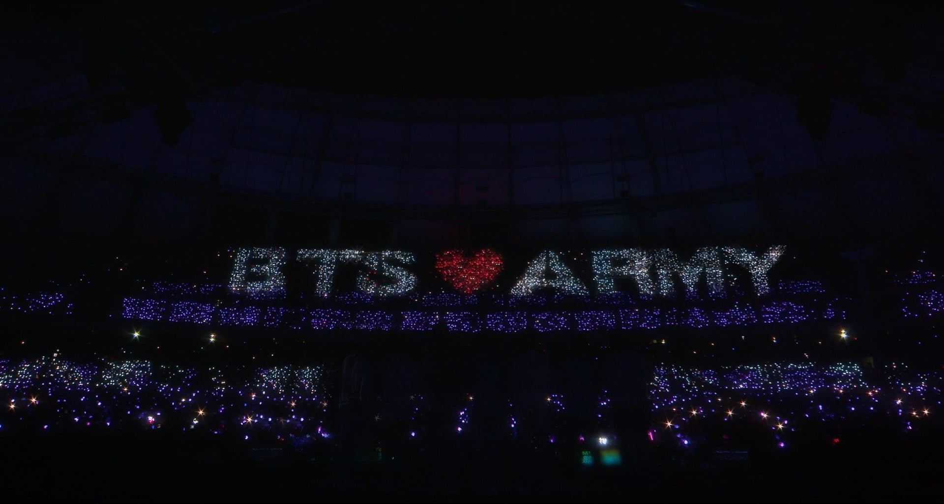 A dark stadium filled with lights spelling out BTS ♥ ARMY above the crowd, with fans holding glowing light sticks throughout the venue.