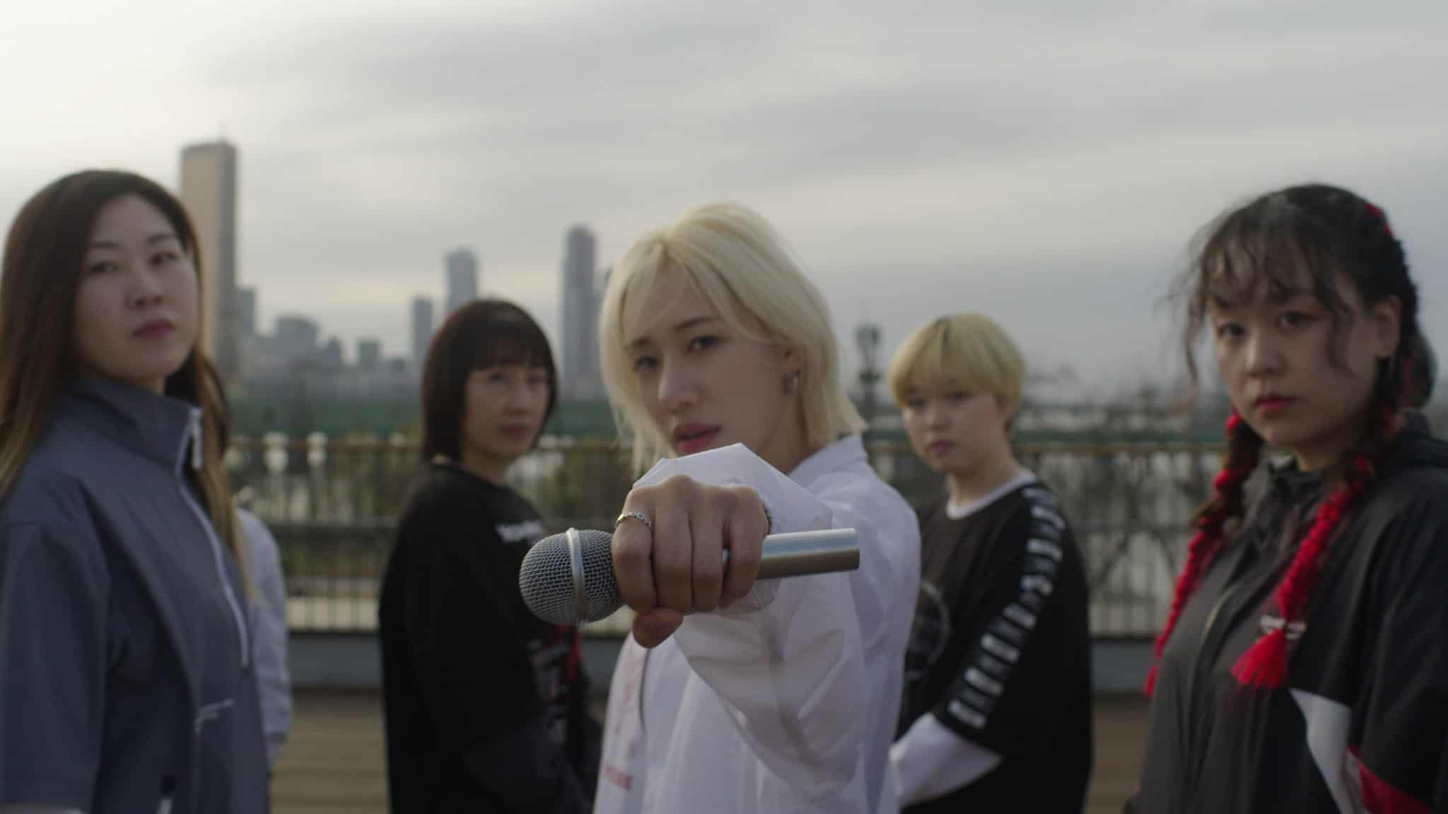 A person with blond hair holds a microphone toward the camera, surrounded by four others, all standing outdoors with a city skyline in the background under a cloudy sky.