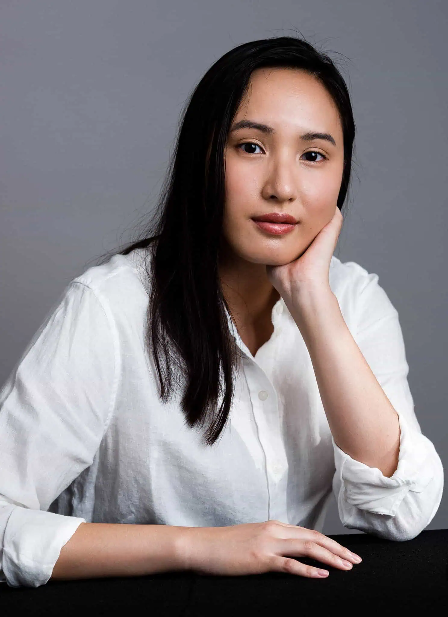 A woman with long dark hair in a white shirt rests her face on her hand next to the book cover for What Hunger by Catherine Dang, which features a close-up of a raspberry and red lips on a black background.