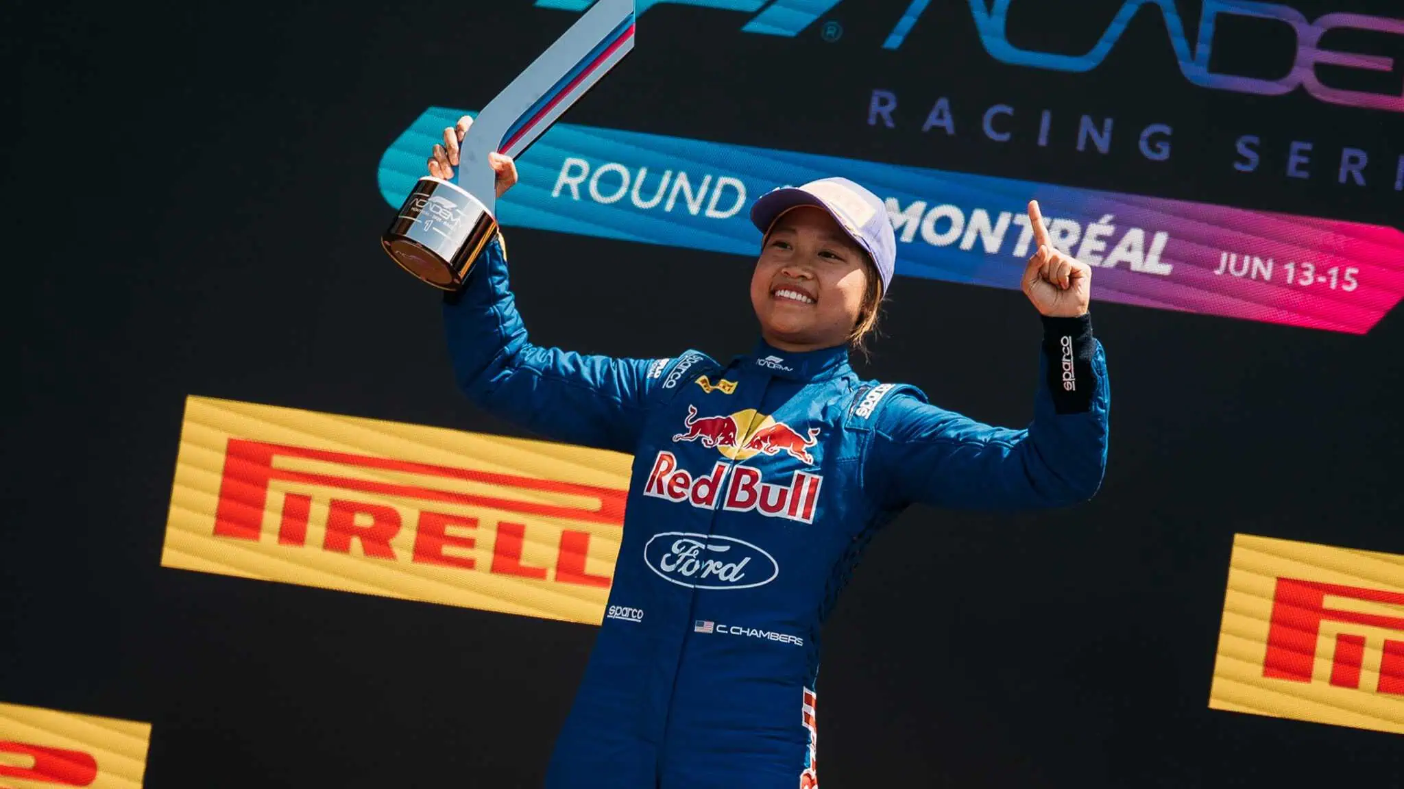 A smiling racing driver in a blue Red Bull Ford suit holds up a trophy and points upward on a podium, with Pirelli and Montreal event signage visible in the background.