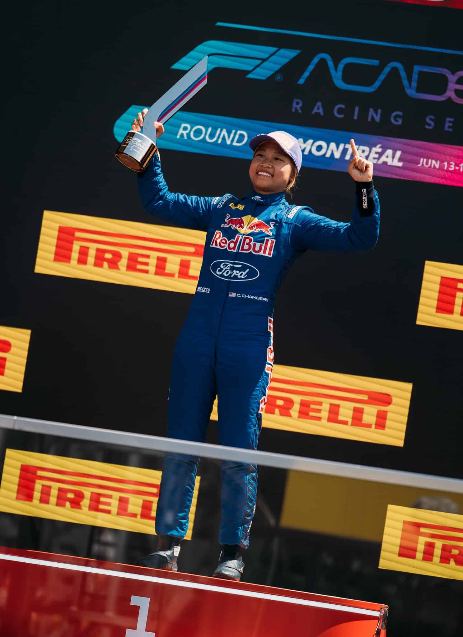 A smiling racing driver in a blue Red Bull Ford suit holds up a trophy and points upward on a podium, with Pirelli and Montreal event signage visible in the background.