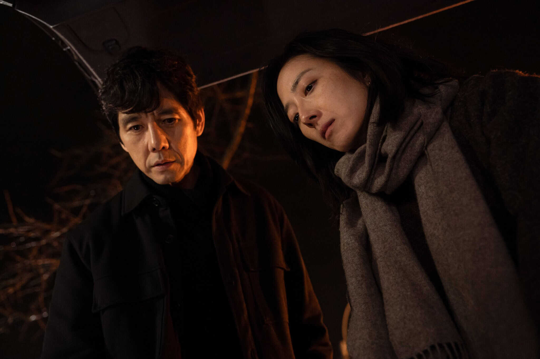 A man and a woman stand close together at night, looking down with serious expressions. The woman wears a scarf and coat, and the man is in a dark jacket. The scene is dimly lit, suggesting a tense or dramatic moment.