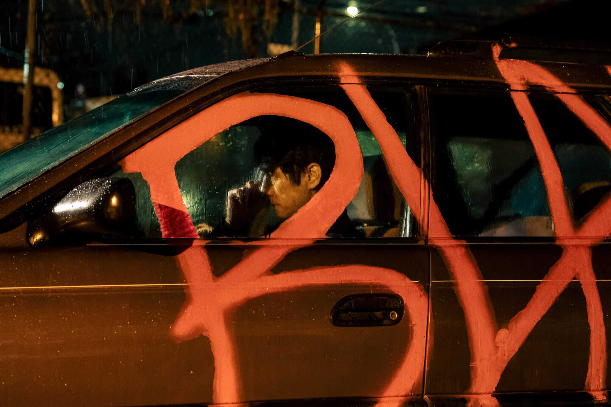 A person sits inside a dark car at night, partially obscured by large red spray-painted letters covering the cars window and door. The person appears thoughtful, touching their face. Rain droplets are visible on the window.
