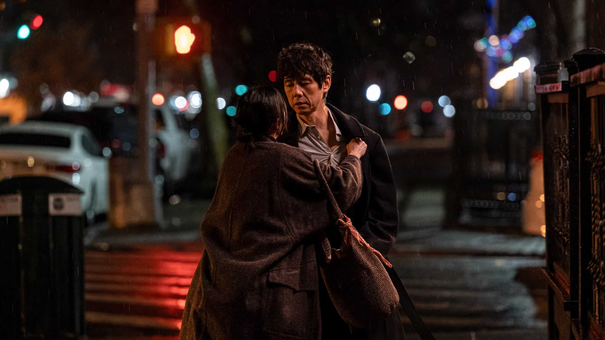 A woman clutches a mans coat under a streetlight at night in the rain. The man looks serious as they stand on a city sidewalk, with blurred car lights and street signs in the background.