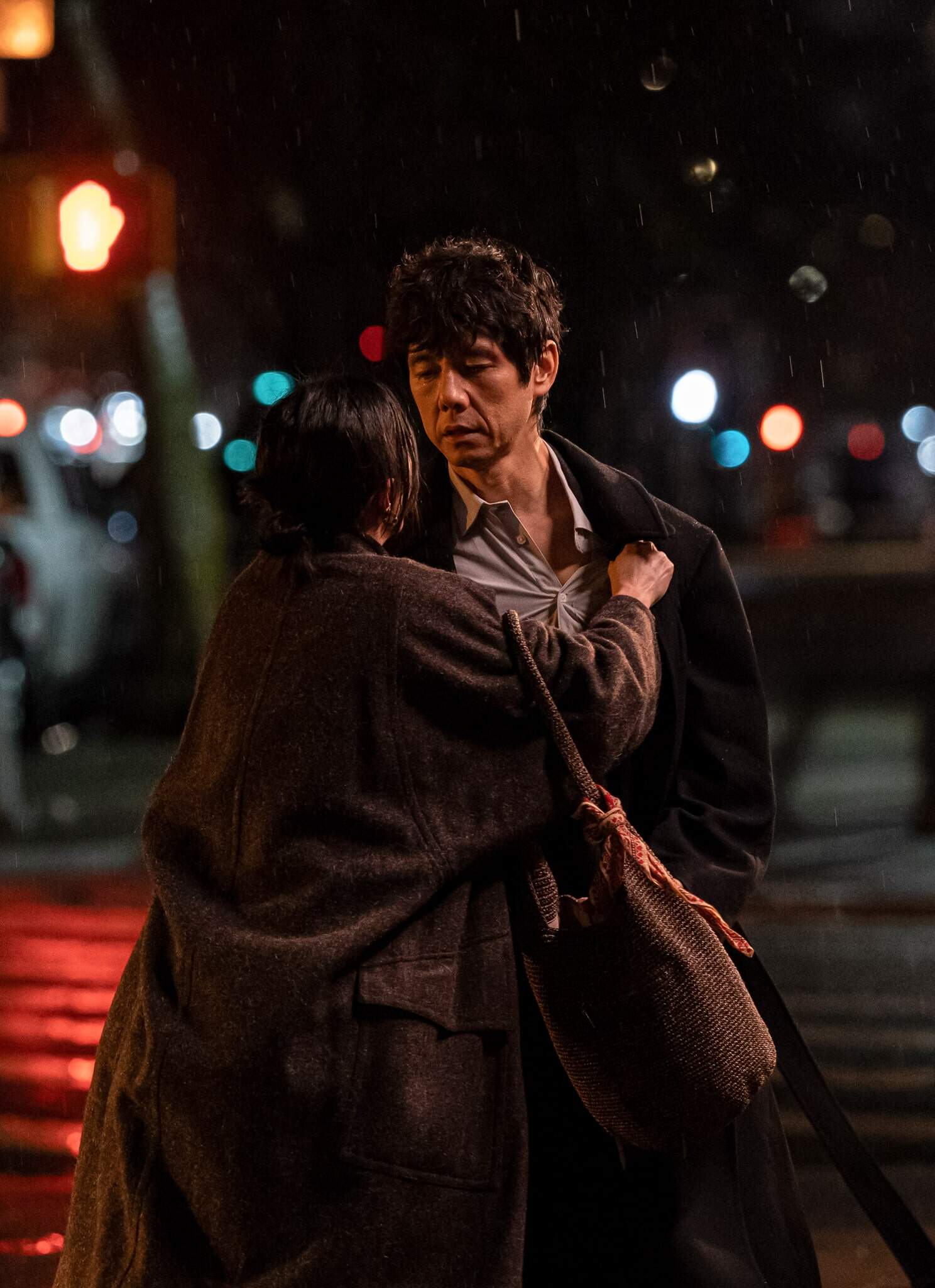 A woman clutches a mans coat under a streetlight at night in the rain. The man looks serious as they stand on a city sidewalk, with blurred car lights and street signs in the background.