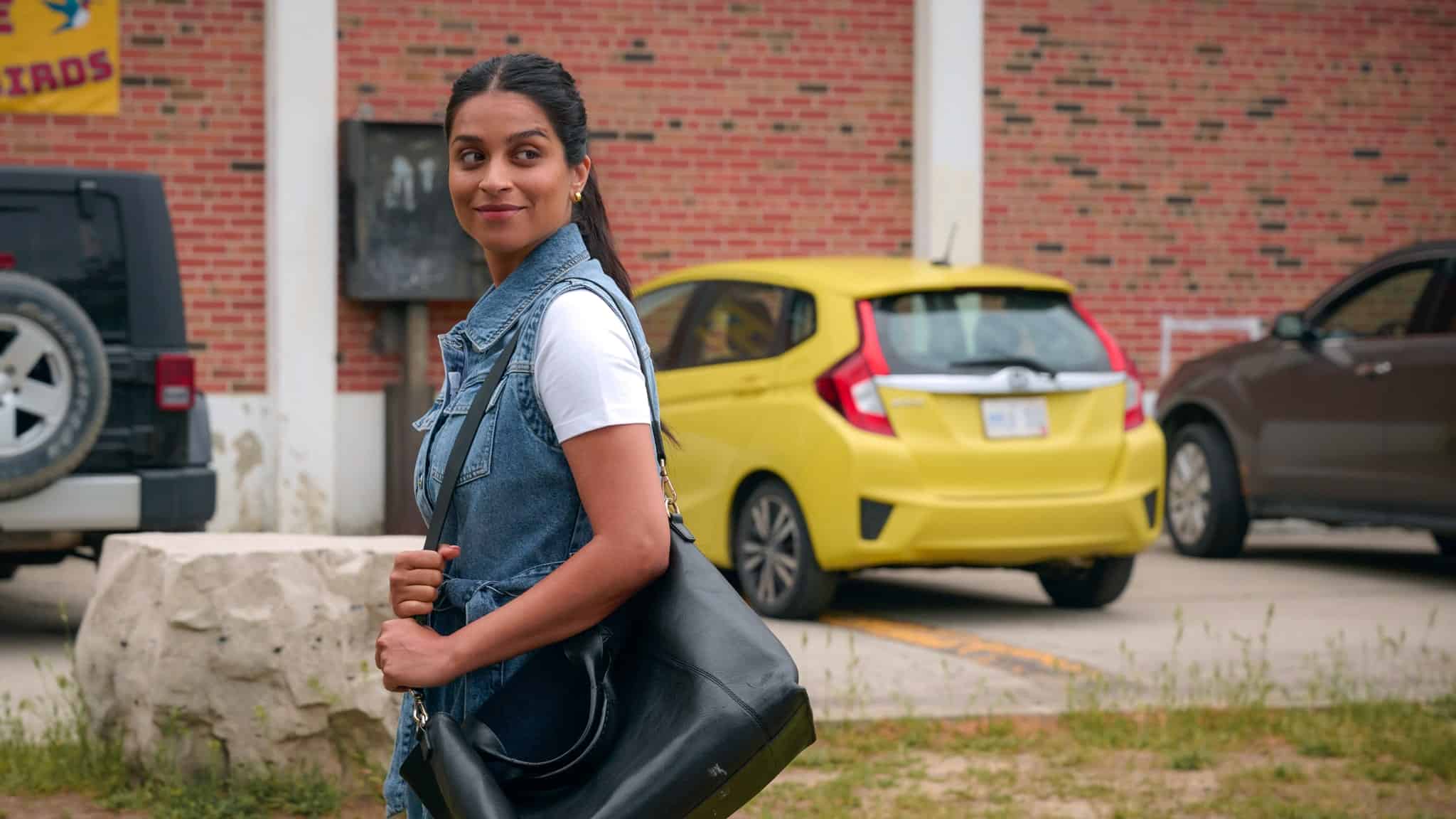 A woman with long dark hair in a ponytail, wearing a white shirt and denim vest, stands outdoors smiling and holding a black bag. Behind her are parked cars, including a yellow hatchback, and a brick building.