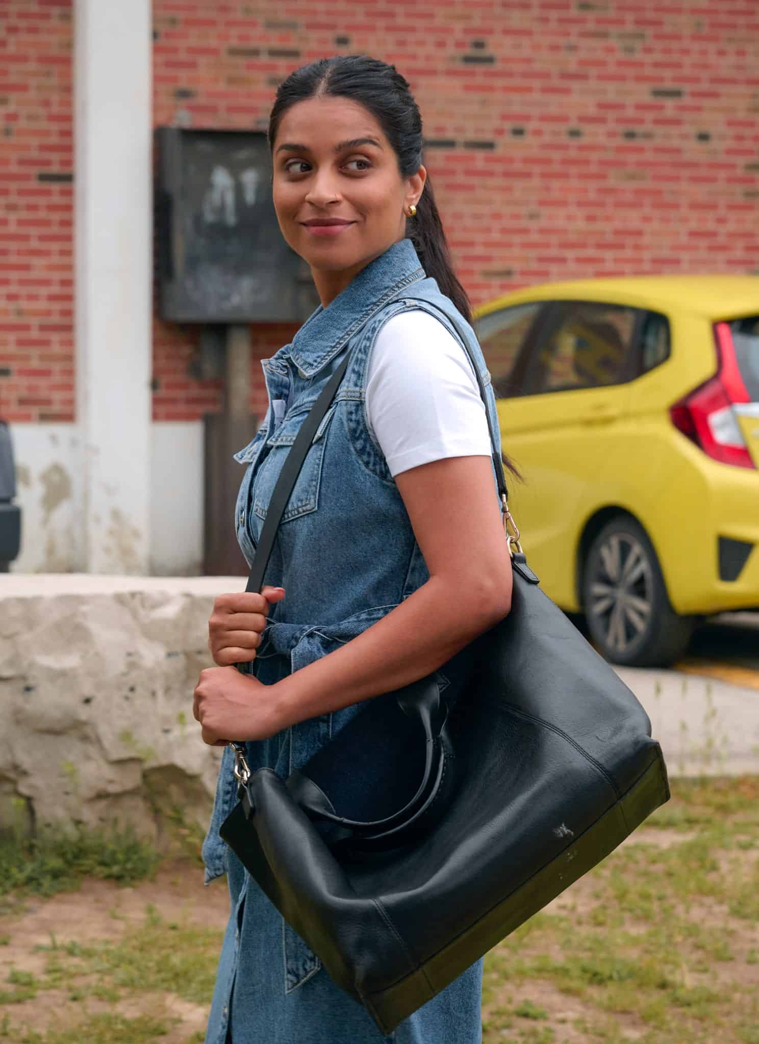 A woman with long dark hair in a ponytail, wearing a white shirt and denim vest, stands outdoors smiling and holding a black bag. Behind her are parked cars, including a yellow hatchback, and a brick building.