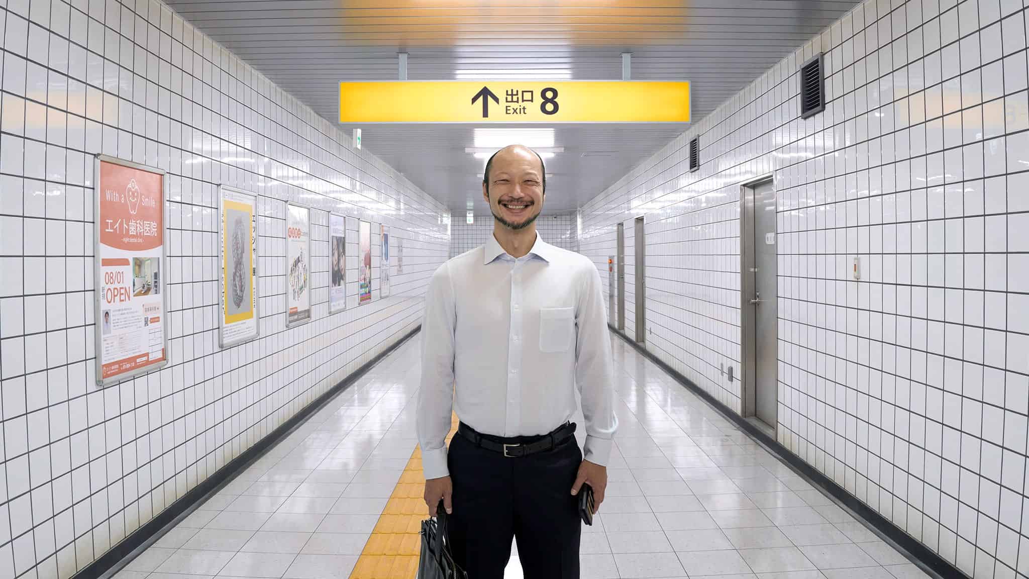 A man in a white shirt stands smiling in a tiled subway corridor beneath a yellow sign that reads Exit 8 in English and Japanese. He is holding a briefcase and facing the camera.