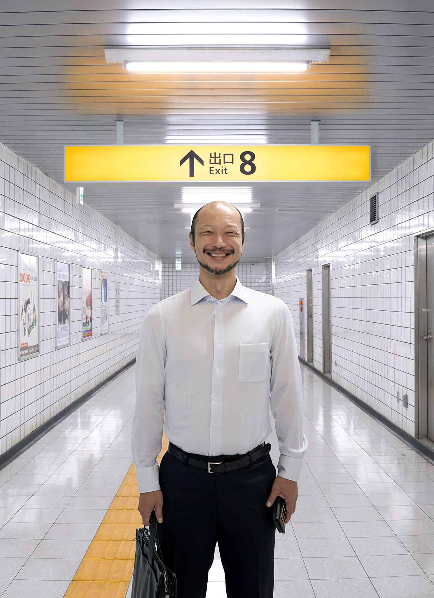 A man in a white shirt stands smiling in a tiled subway corridor beneath a yellow sign that reads Exit 8 in English and Japanese. He is holding a briefcase and facing the camera.