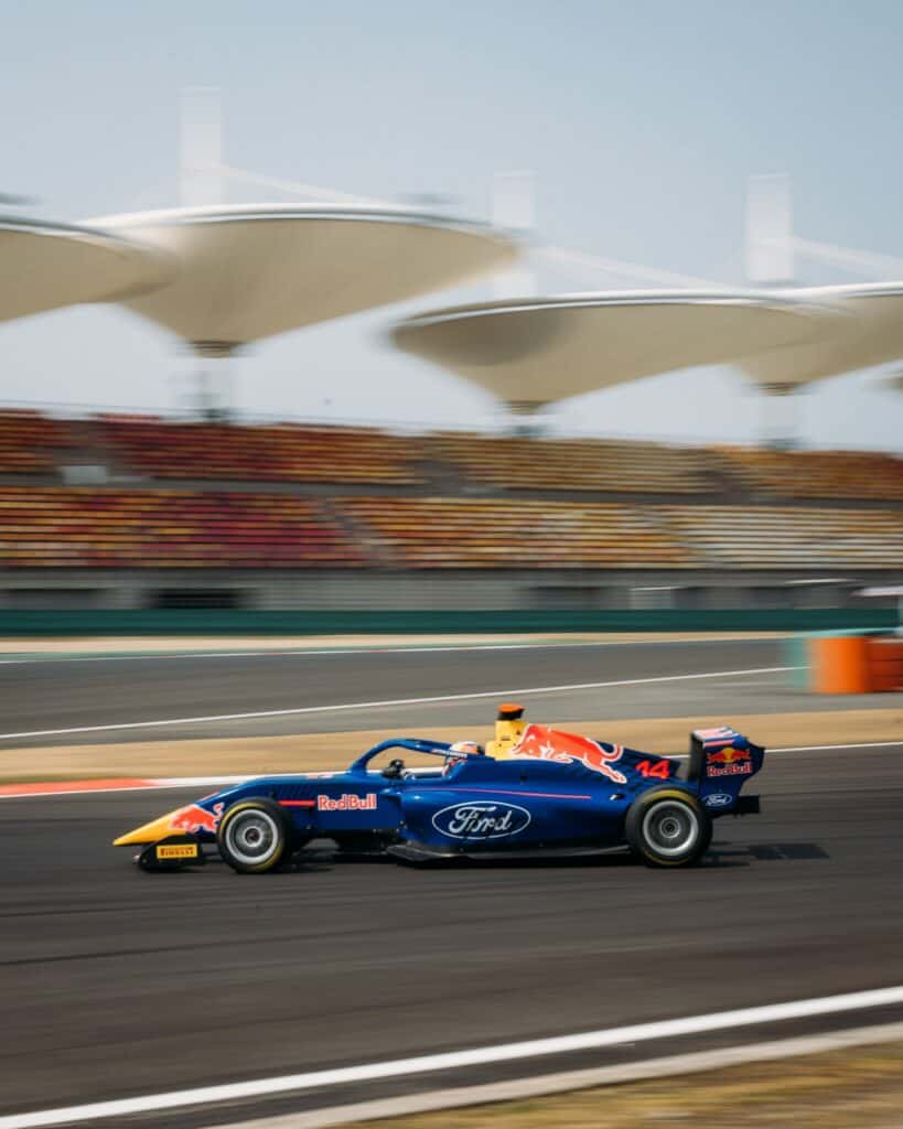 A blue and yellow Formula 3 race car with Red Bull and Ford logos speeds along a racetrack, with blurred grandstands and canopied structures in the background.
