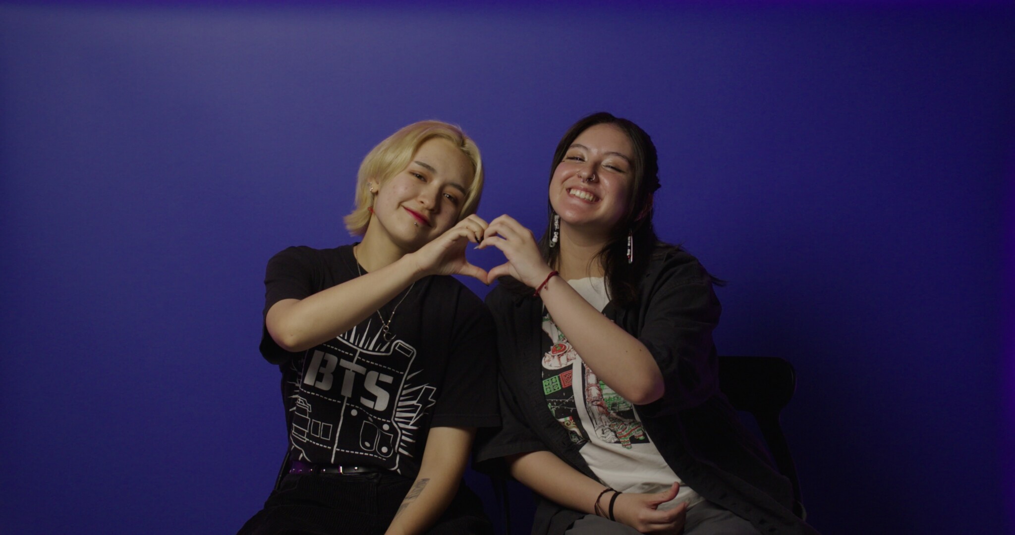 Two people sit together against a blue background, smiling and forming a heart shape with their hands. One wears a black BTS shirt, and the other wears a white and gray shirt with a graphic print.