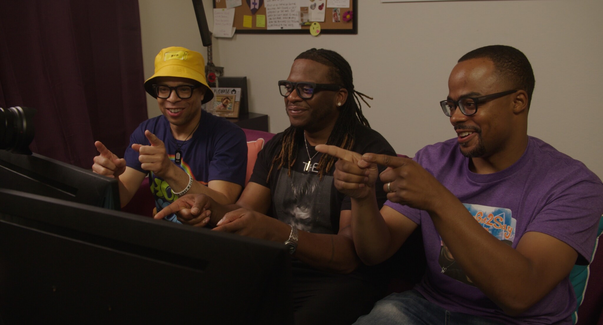 Three men sitting side by side smile and point at computer monitors in front of them. They all wear glasses and casual clothes, and a bulletin board is visible in the background.