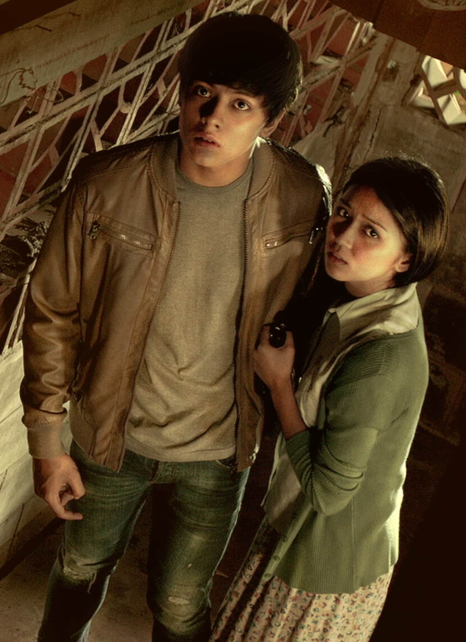 A young man and woman stand close together indoors, looking up with anxious expressions. The lighting and setting create a tense, suspenseful mood. The man wears a brown jacket and jeans; the woman wears a green cardigan and floral skirt.