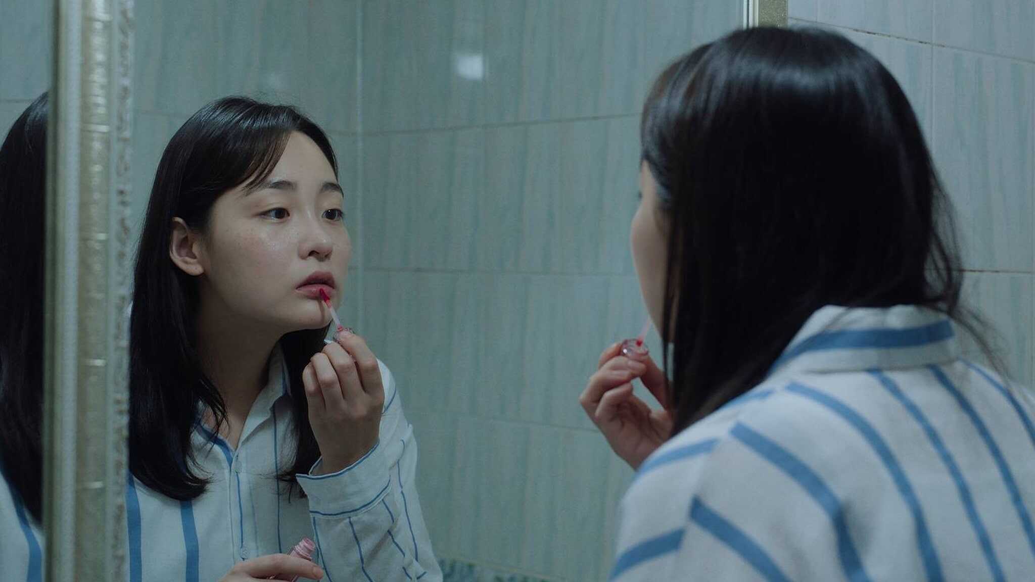 A woman wearing a blue-and-white striped pajama top applies lipstick while looking at herself in a bathroom mirror with tiled walls.