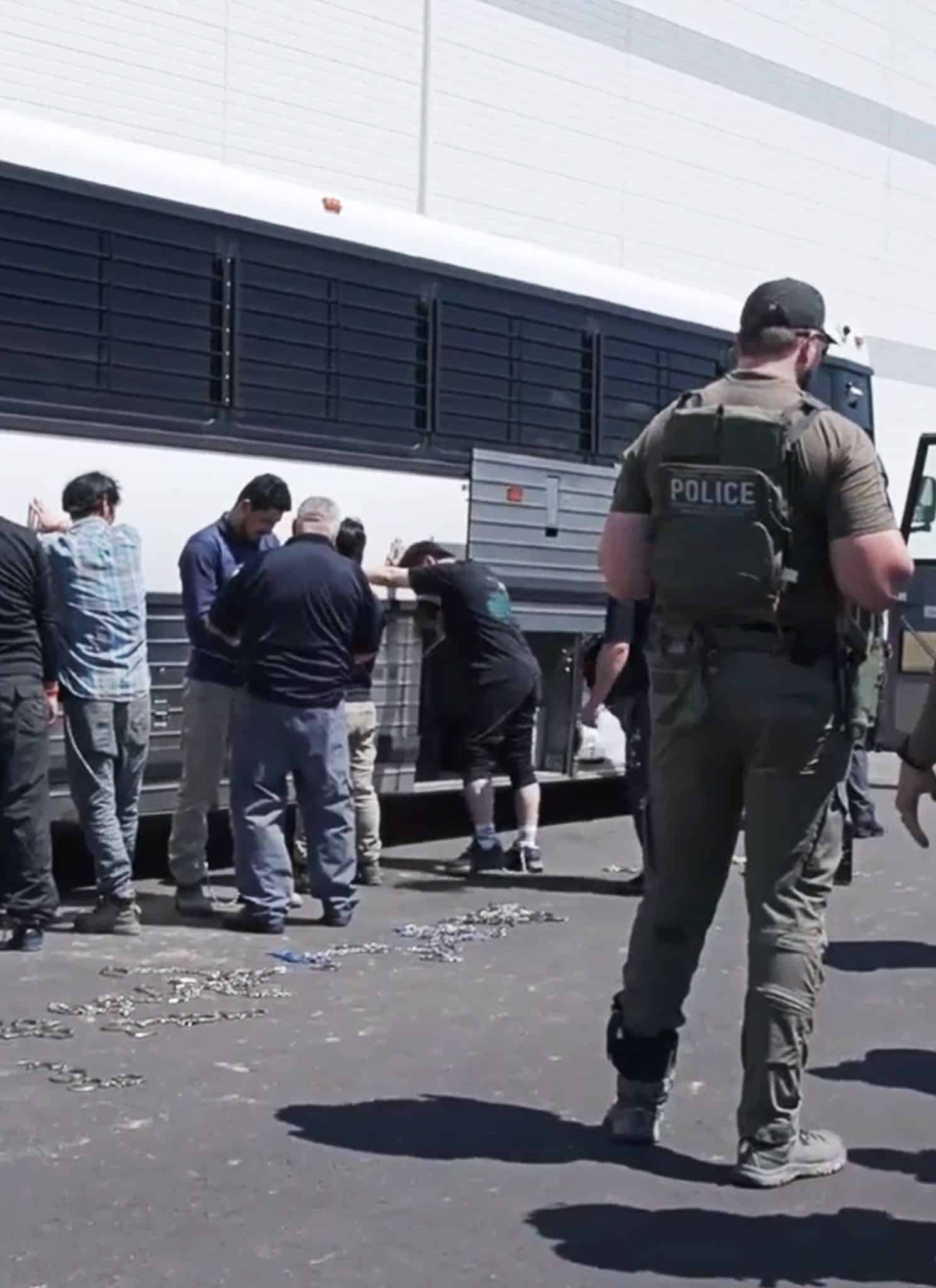 A group of people stand facing a bus with their hands against it while law enforcement officers in tactical gear stand nearby, monitoring the scene outdoors on a paved surface.