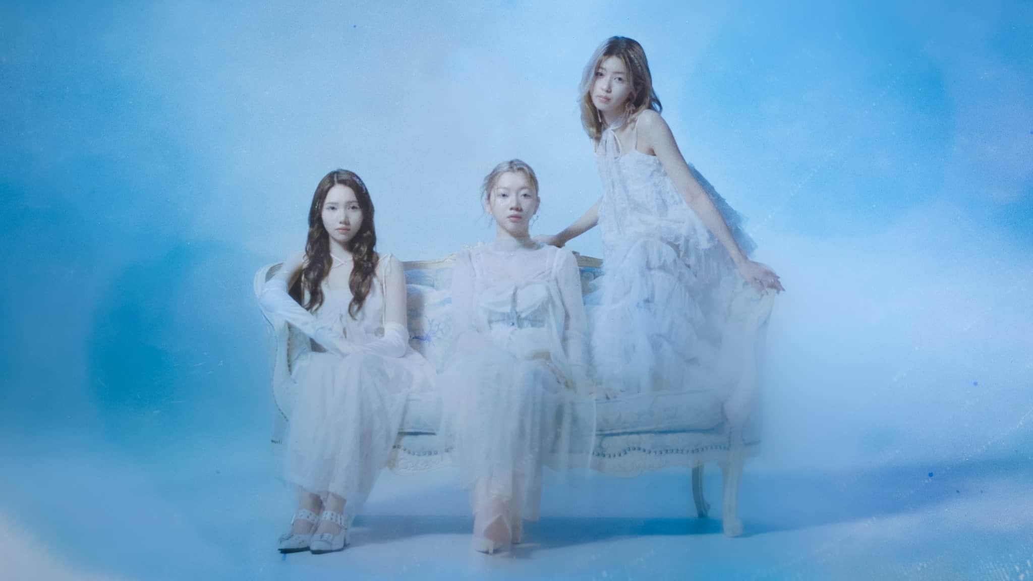 Three young women in long, white, elegant dresses sit and pose on a vintage blue and white sofa, surrounded by a soft, dreamy blue background. The scene has an ethereal and serene atmosphere.