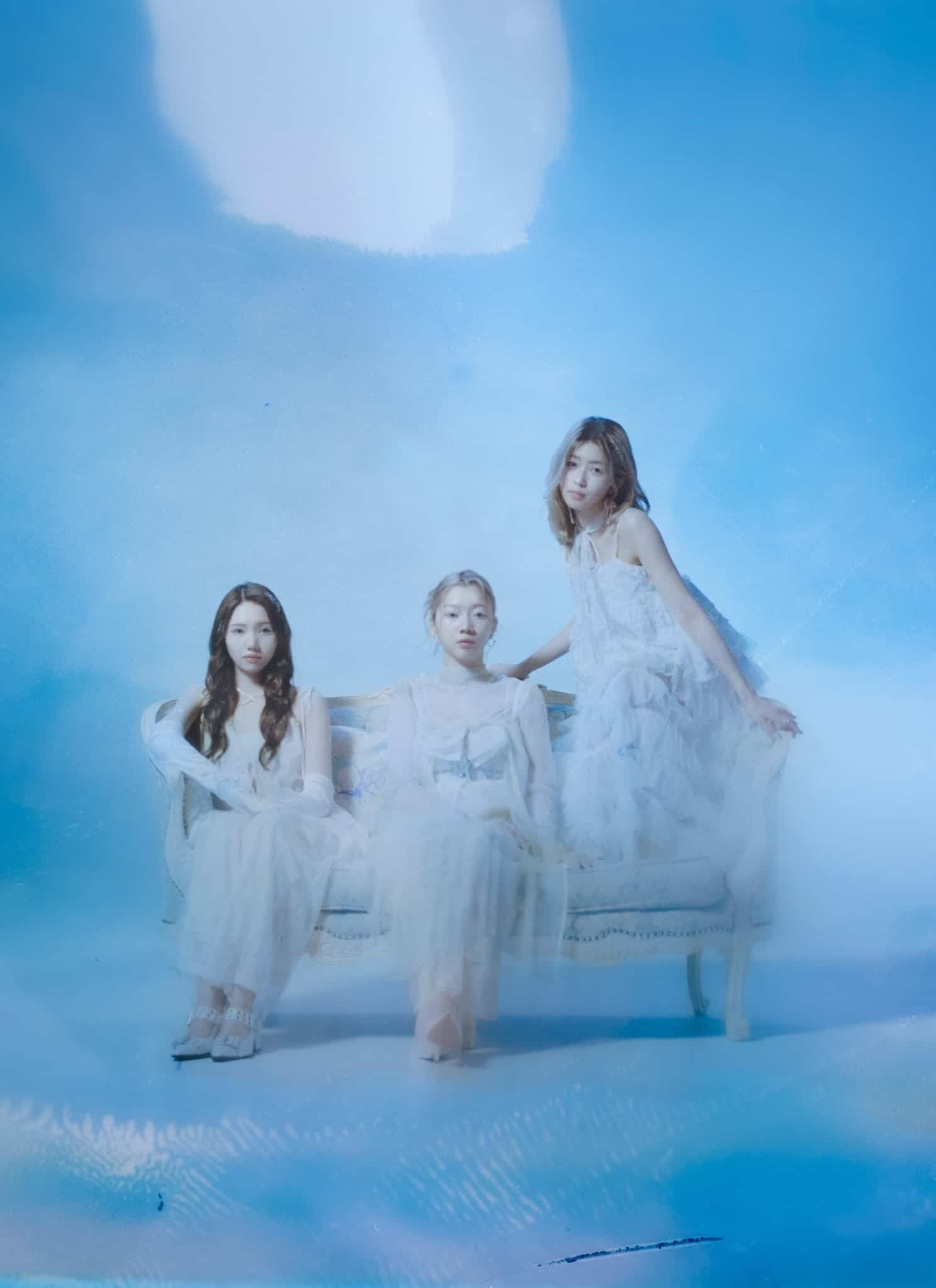 Three young women in long, white, elegant dresses sit and pose on a vintage blue and white sofa, surrounded by a soft, dreamy blue background. The scene has an ethereal and serene atmosphere.